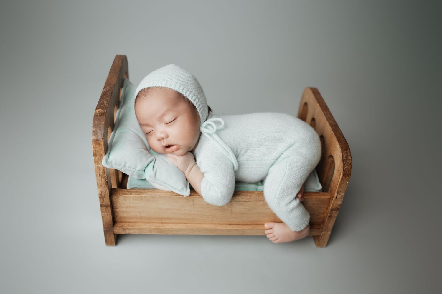 Sleeping newborn in mint green romper and bonnet resting on wooden toy bed with pillow.