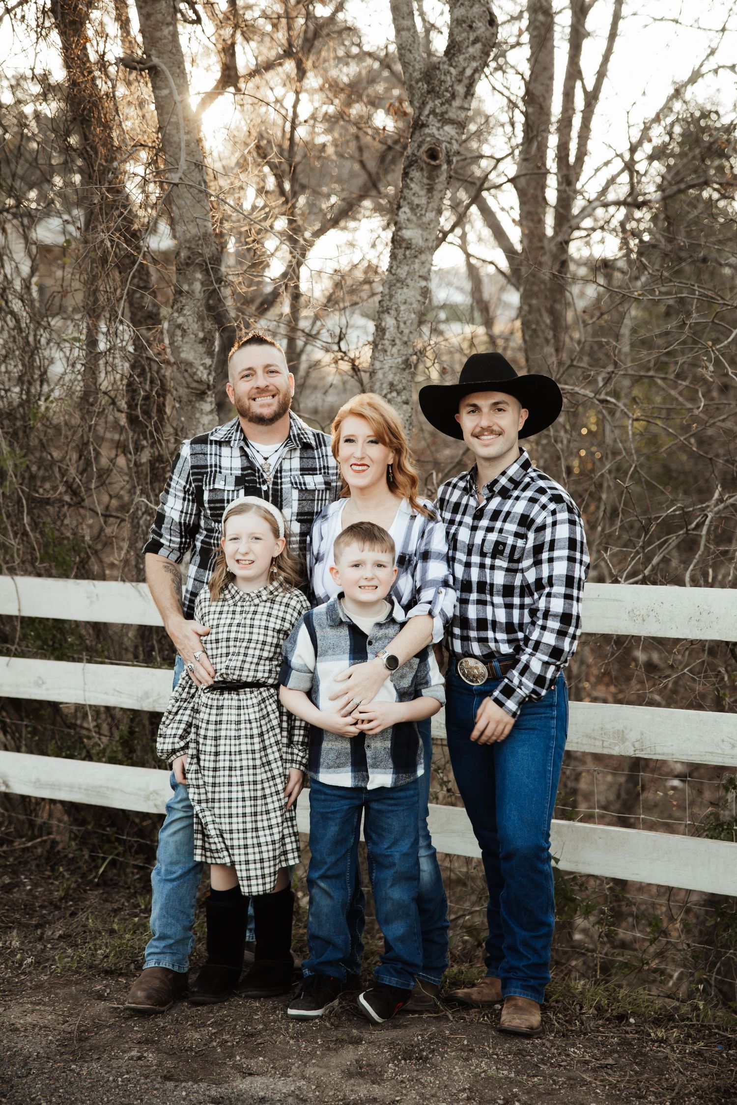 A family of five in matching plaid outfits poses by a white fence in a wooded area.
