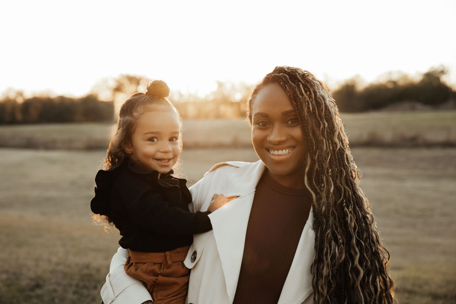 A smiling Black mother with long braids holds her young daughter in a sunlit field.