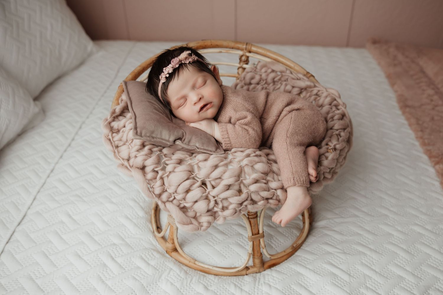 Sleeping newborn baby girl in pink knit outfit rests peacefully in a round rattan basket with fluffy blanket.