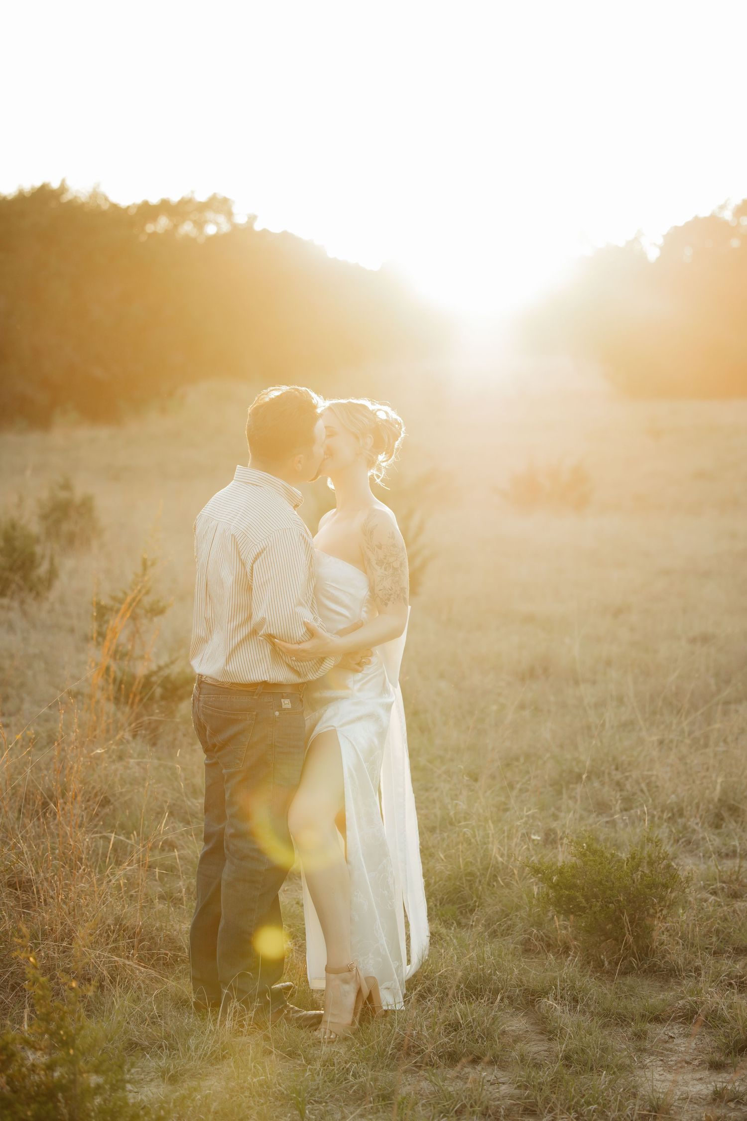 Couple shares romantic kiss bathed in warm golden sunset light in rustic field during wedding photo shoot.