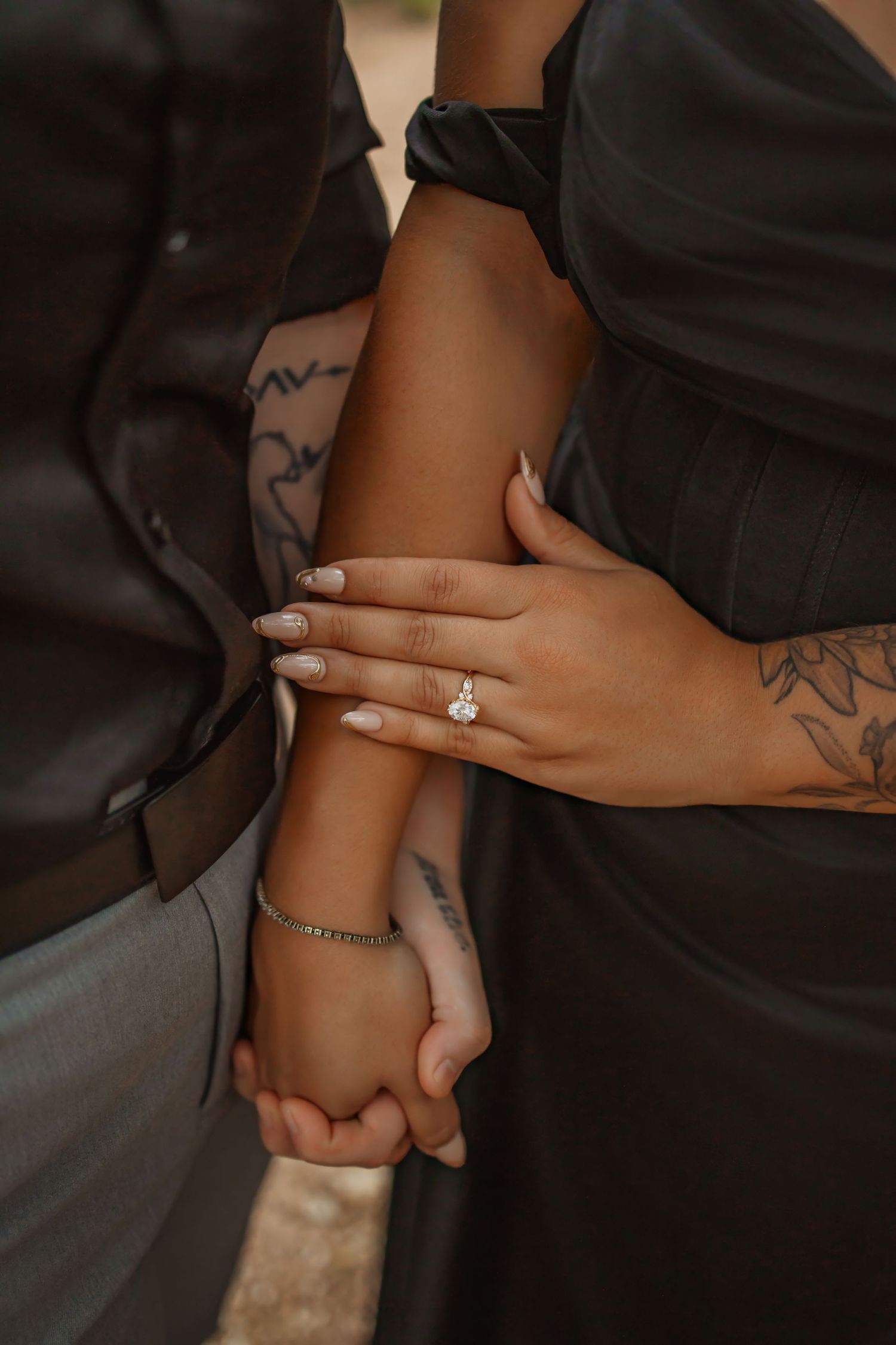 Couple holding hands showing off a diamond engagement ring, with tattoos and neutral manicure visible.