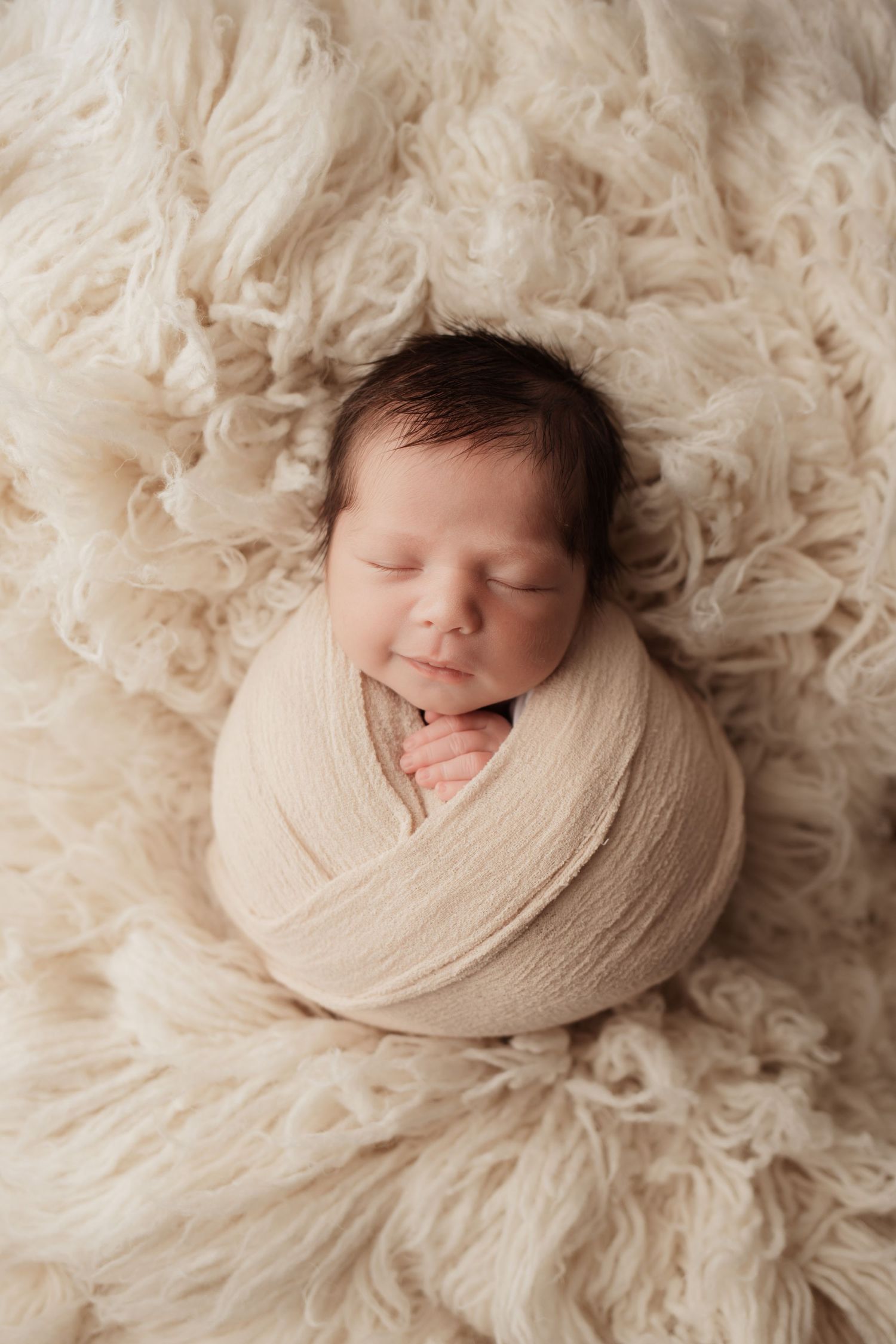 Smiling newborn baby swaddled in cream wrap, sleeping on a soft fluffy ivory flokati rug.