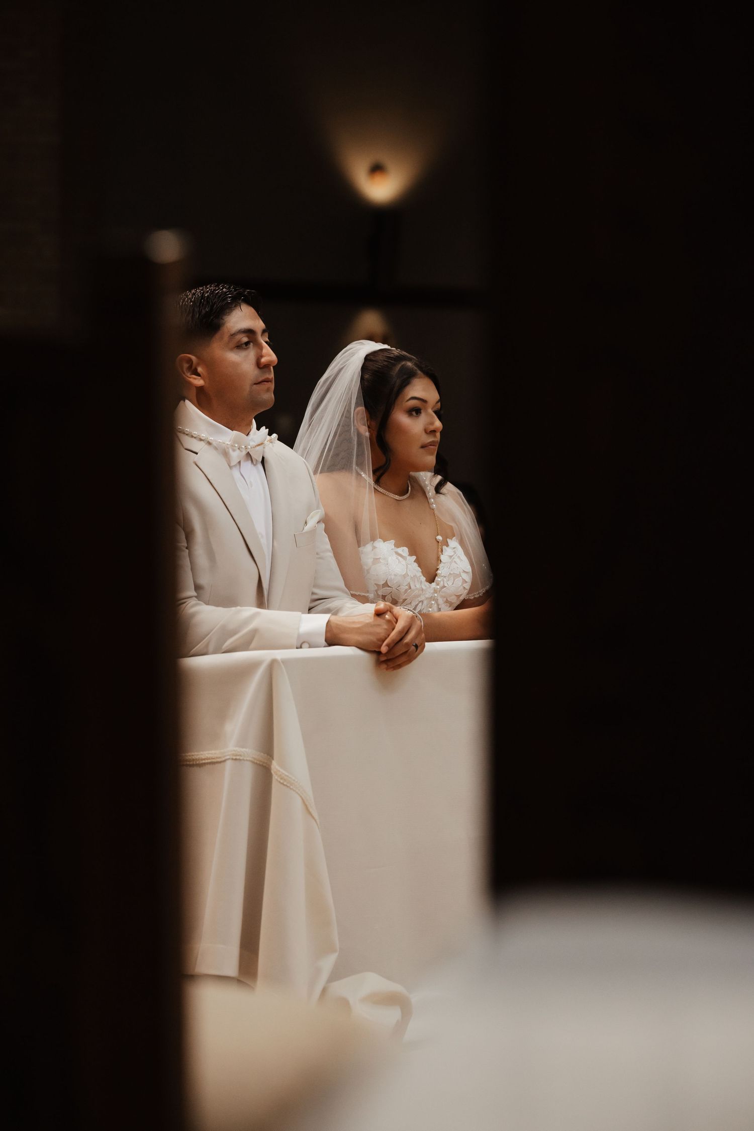 Bride and groom kneel at the altar during a church wedding ceremony, dressed in white gown and ivory suit.