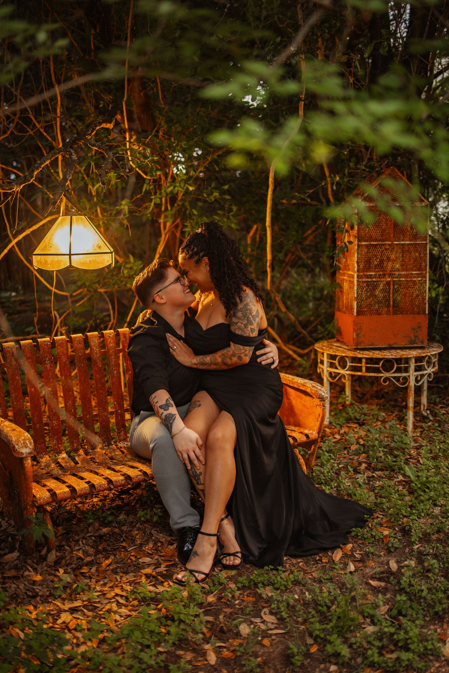 Couple in black attire share an intimate moment on a vintage bench surrounded by warm lantern light outdoors.
