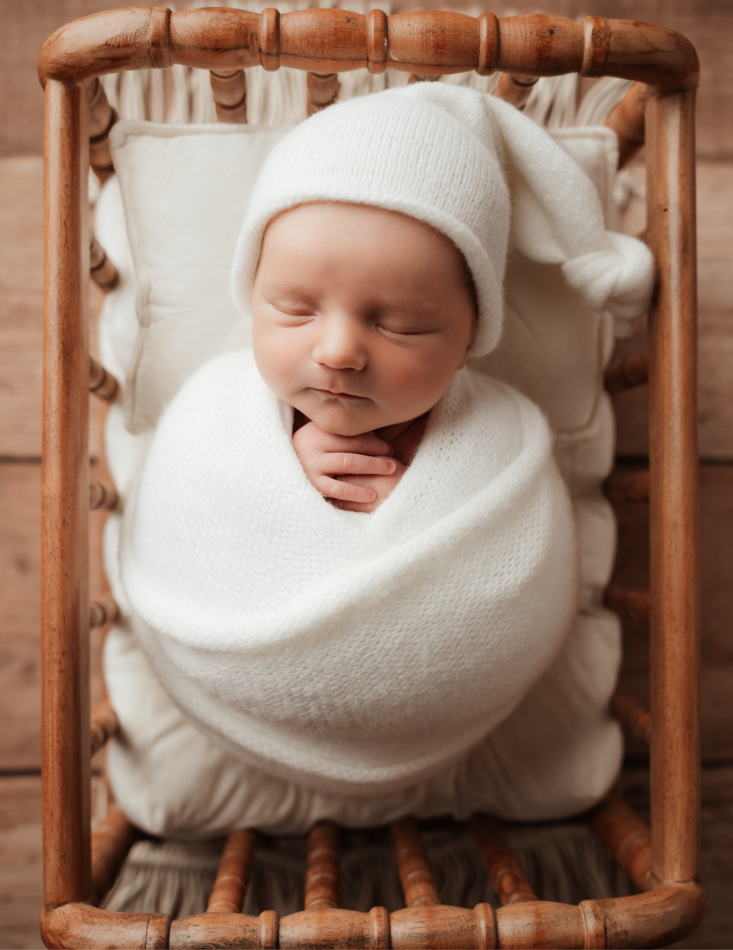 Sleeping newborn baby swaddled in white wrap wearing knit hat in wooden rattan crib prop.