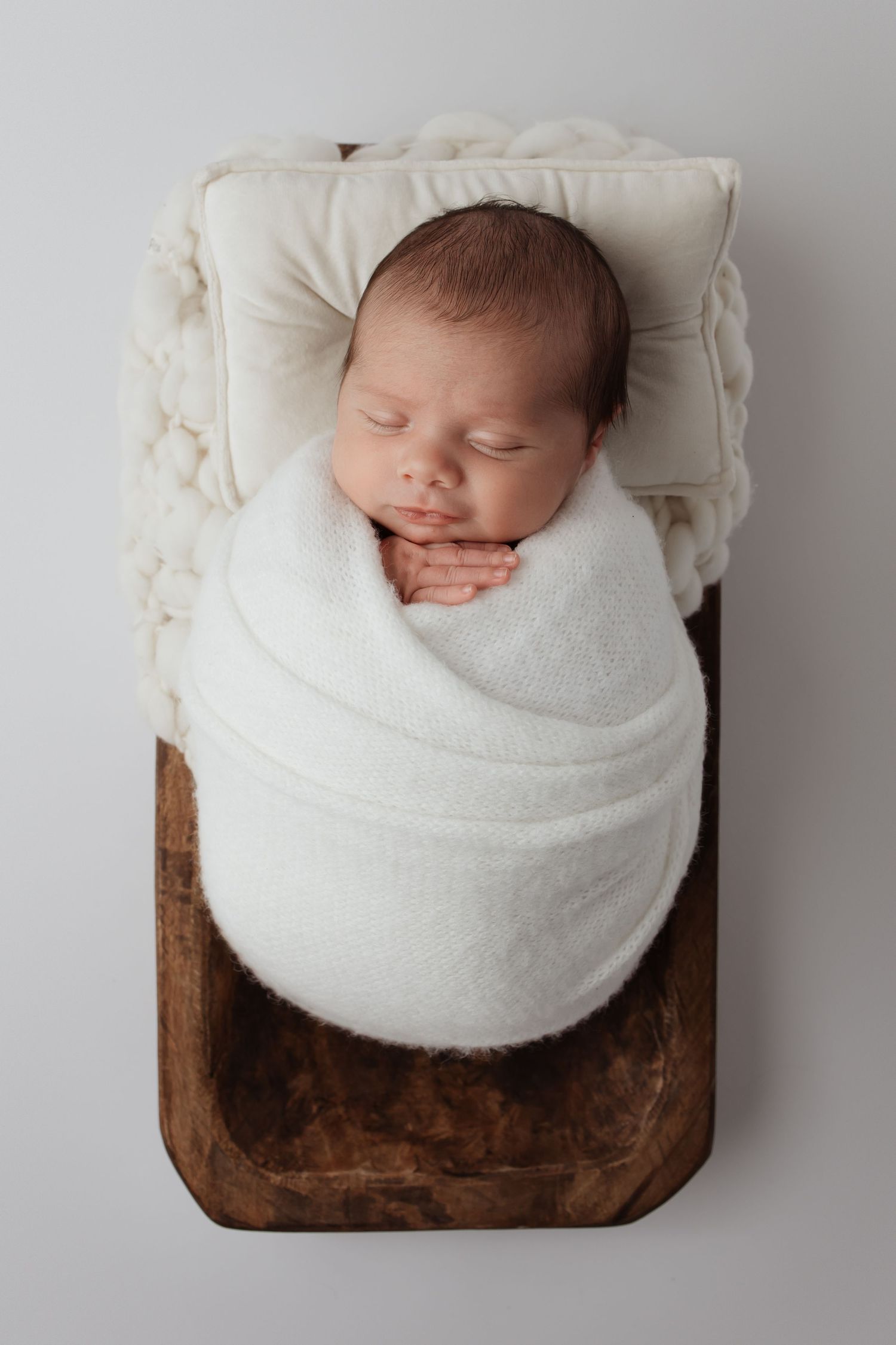 Newborn baby swaddled in white wrap sleeping on a small wooden bed with a tiny pillow.