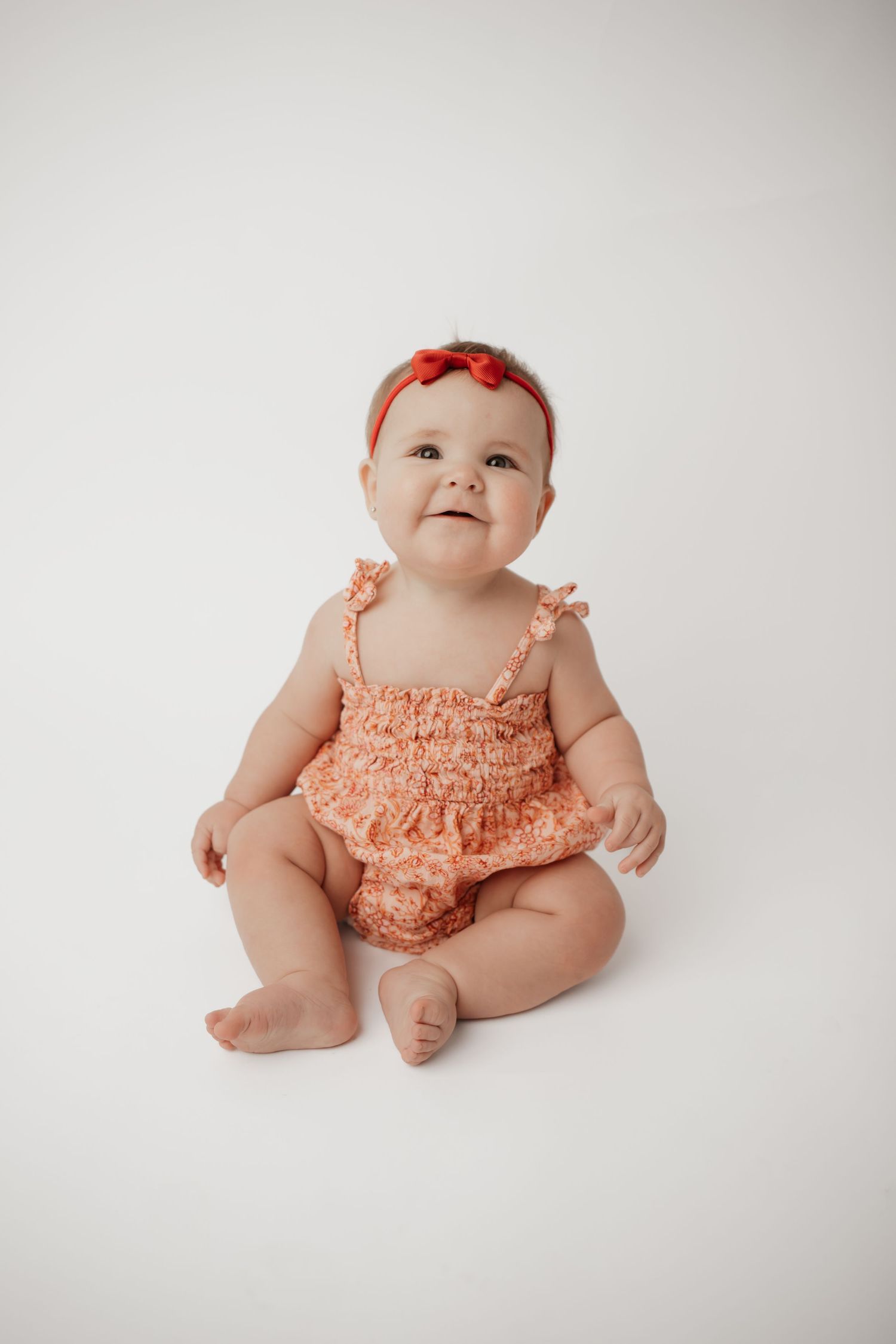 Cute infant in peach floral romper with red headband bow seated on a white studio backdrop.