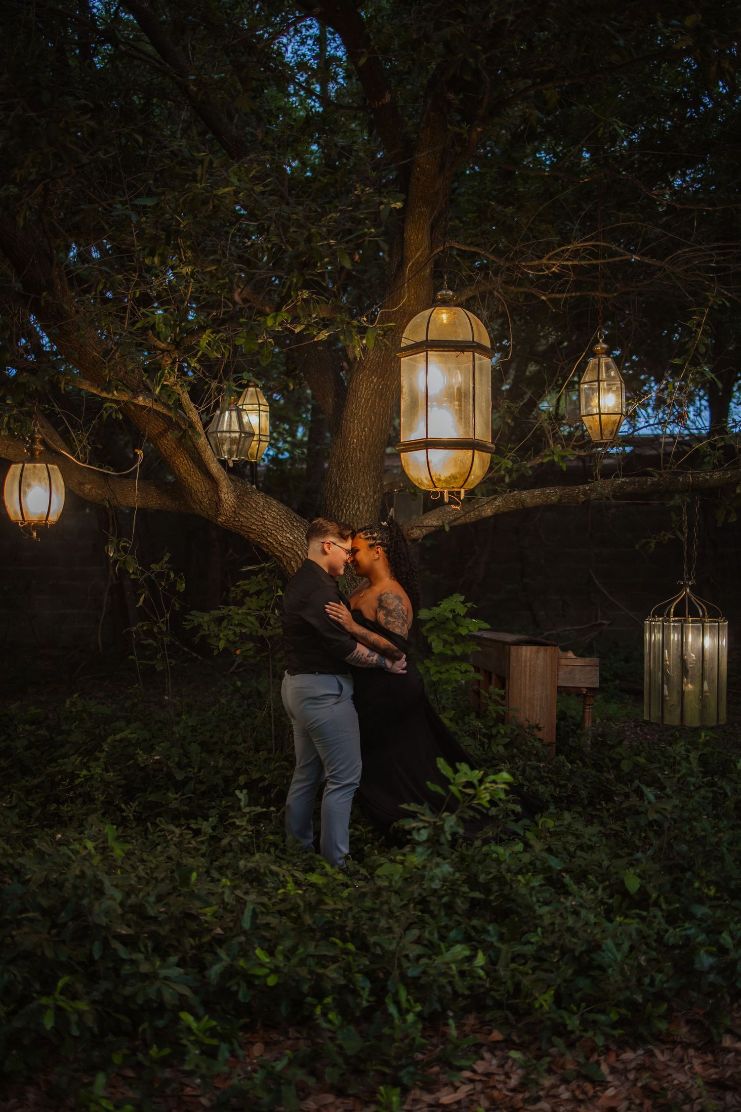 Couple kissing under glowing lanterns hanging from a large tree in a romantic garden at dusk.
