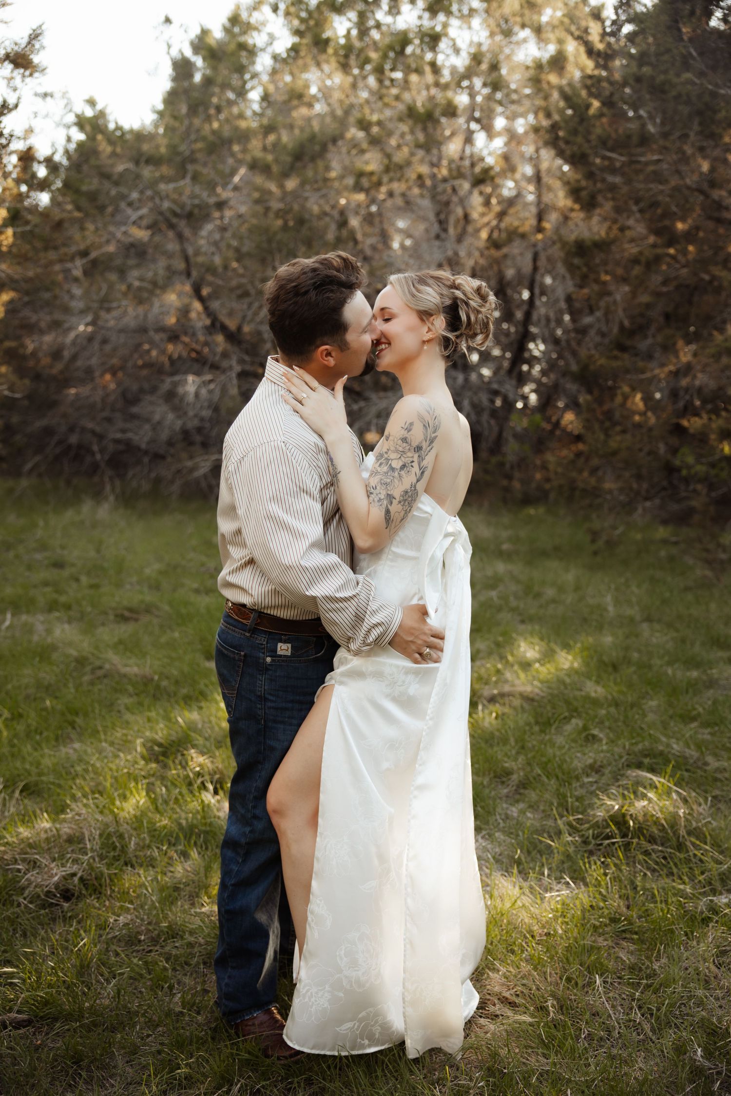 Couple sharing a romantic kiss in a rustic outdoor setting, bride in elegant white gown with tattoos, groom in casual attire.
