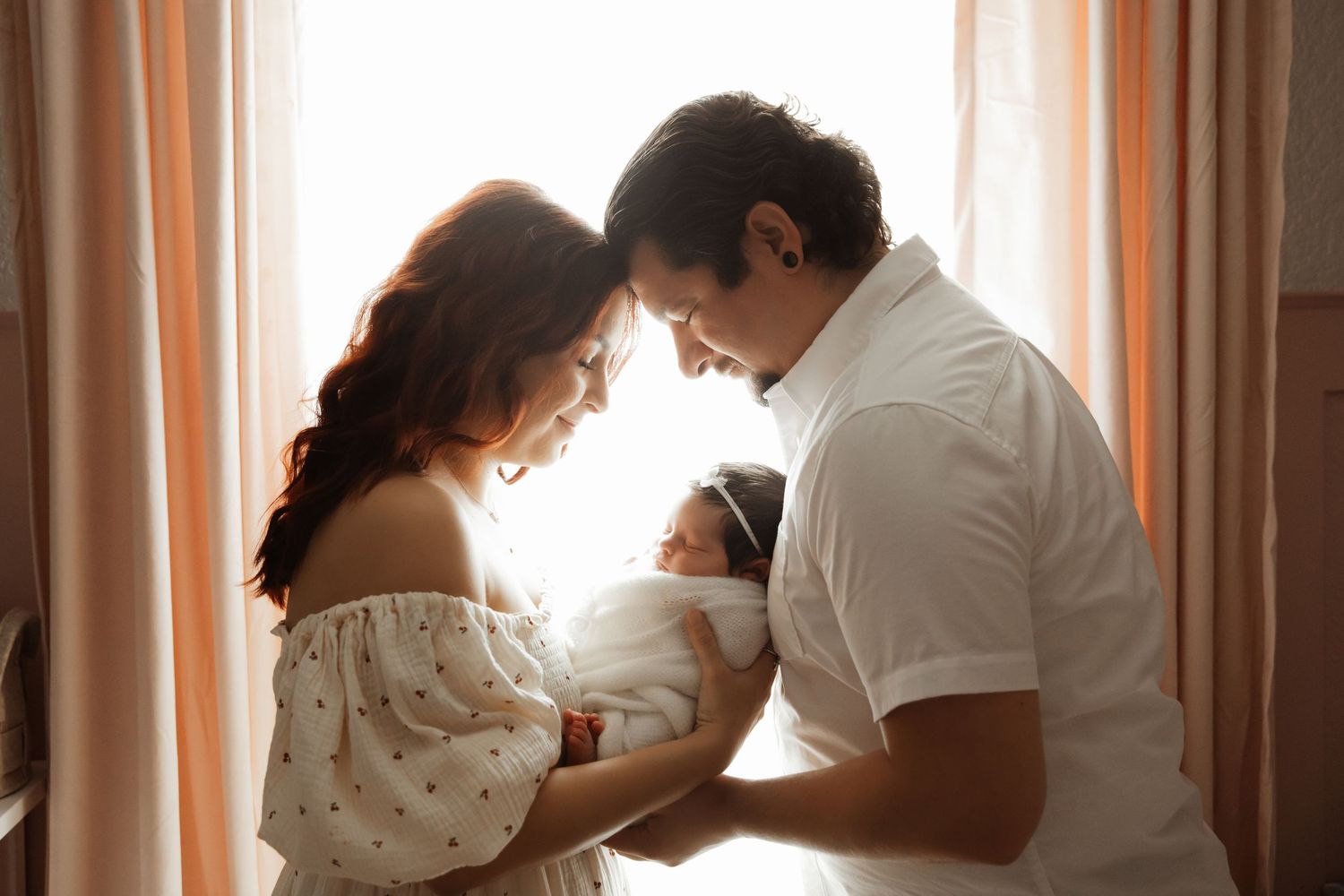New parents tenderly gaze at their newborn baby girl by a sunlit window in a heartwarming family portrait.