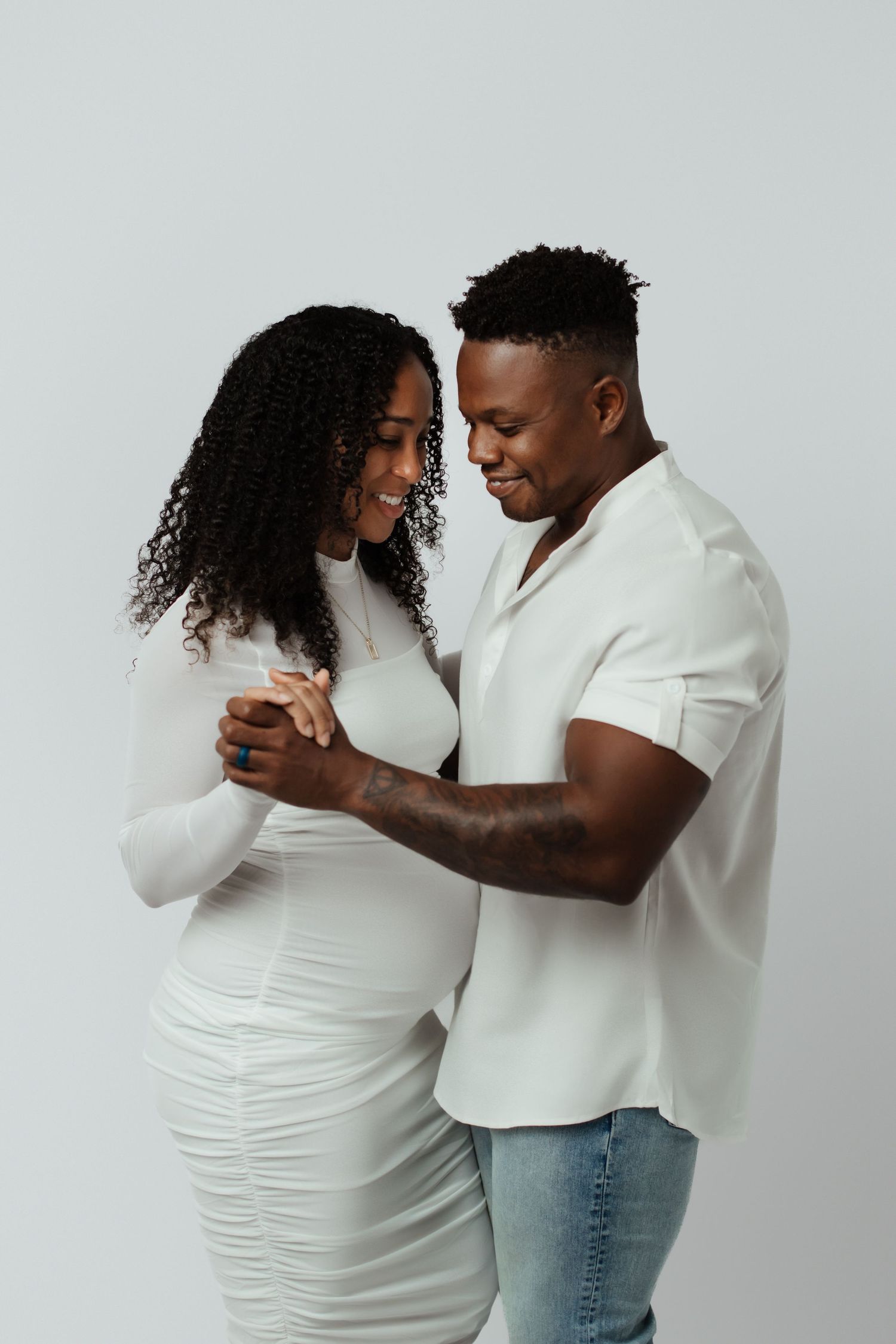 A couple in white outfits holding hands and smiling tenderly at each other against a light gray background.