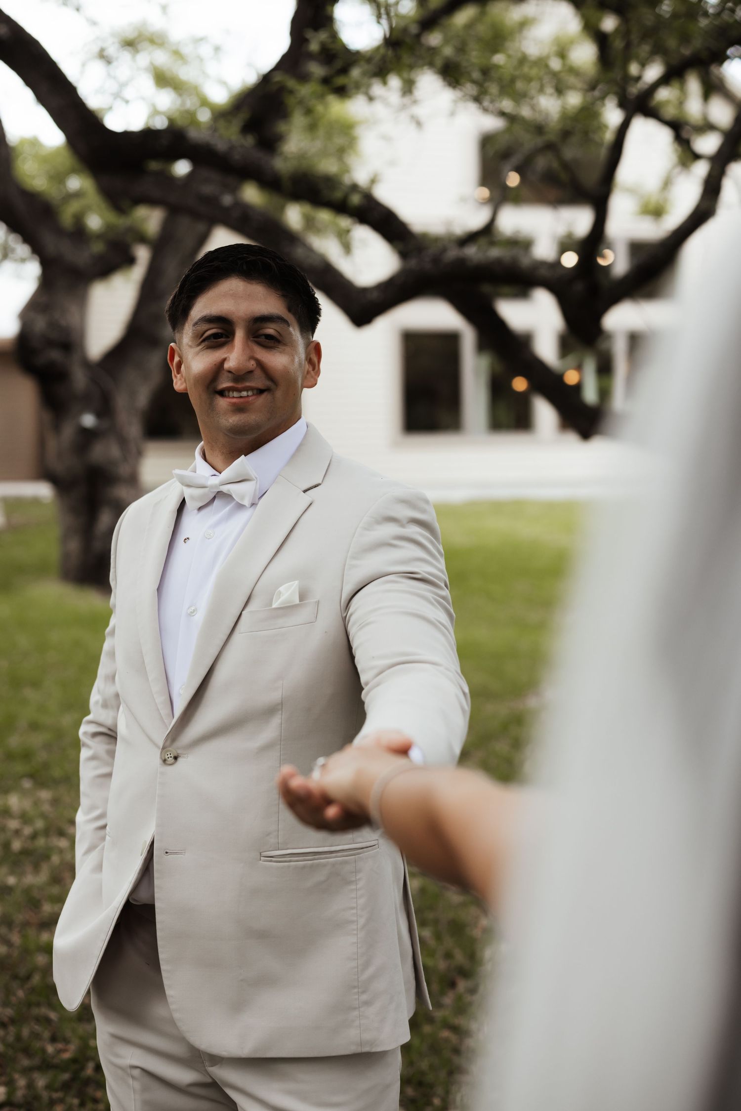 Smiling groom in beige suit and bow tie holds bride's hand outdoors near a large tree at a wedding.