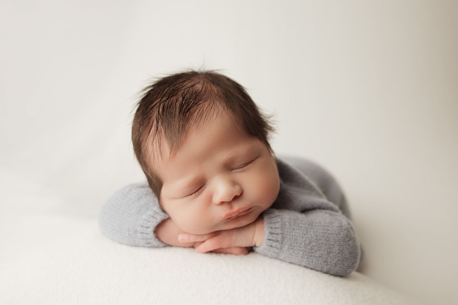 Sleeping newborn baby in gray knit outfit posed with hands under chin on white blanket.