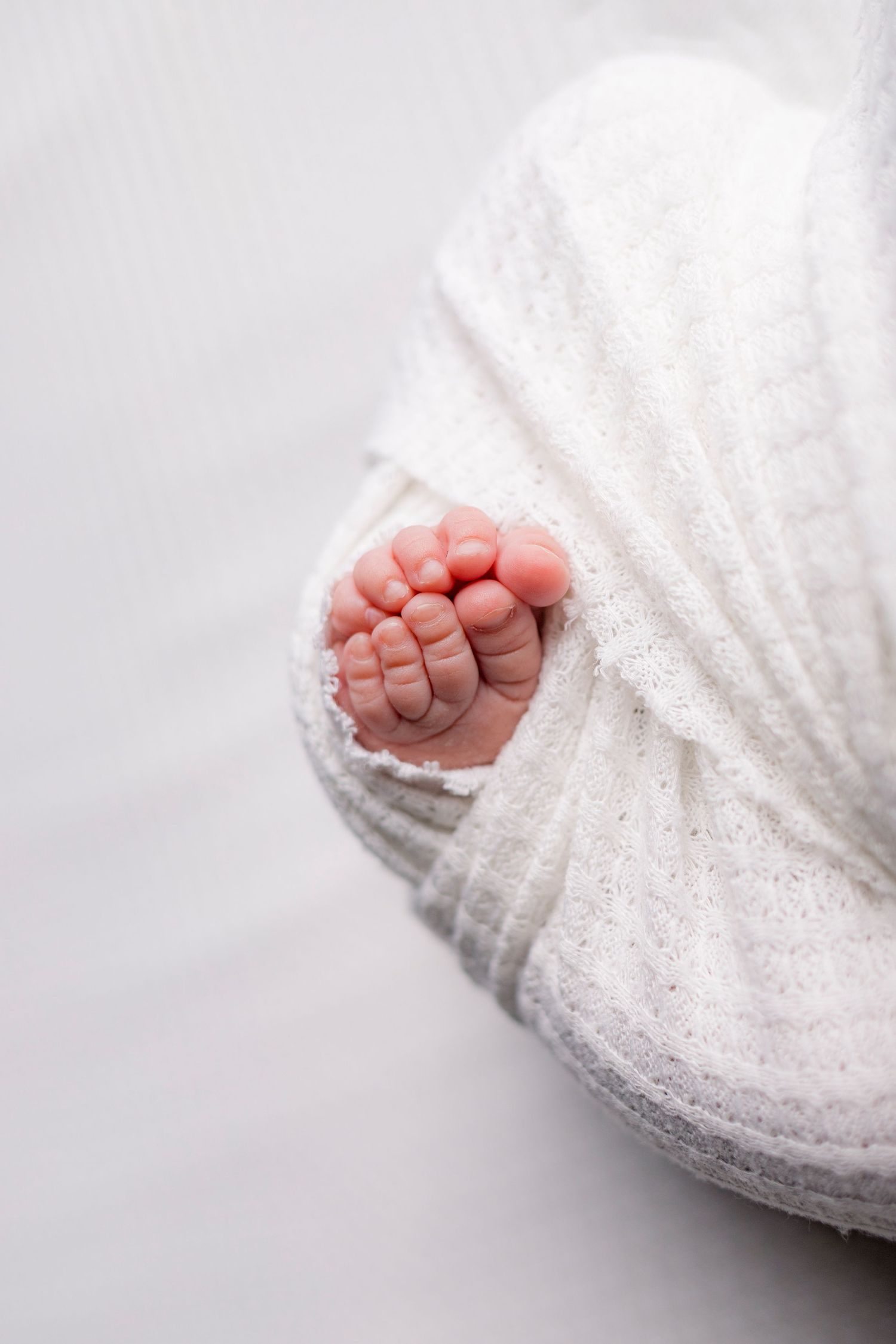 Tiny newborn baby toes peeking out from a soft white knit swaddle wrap.