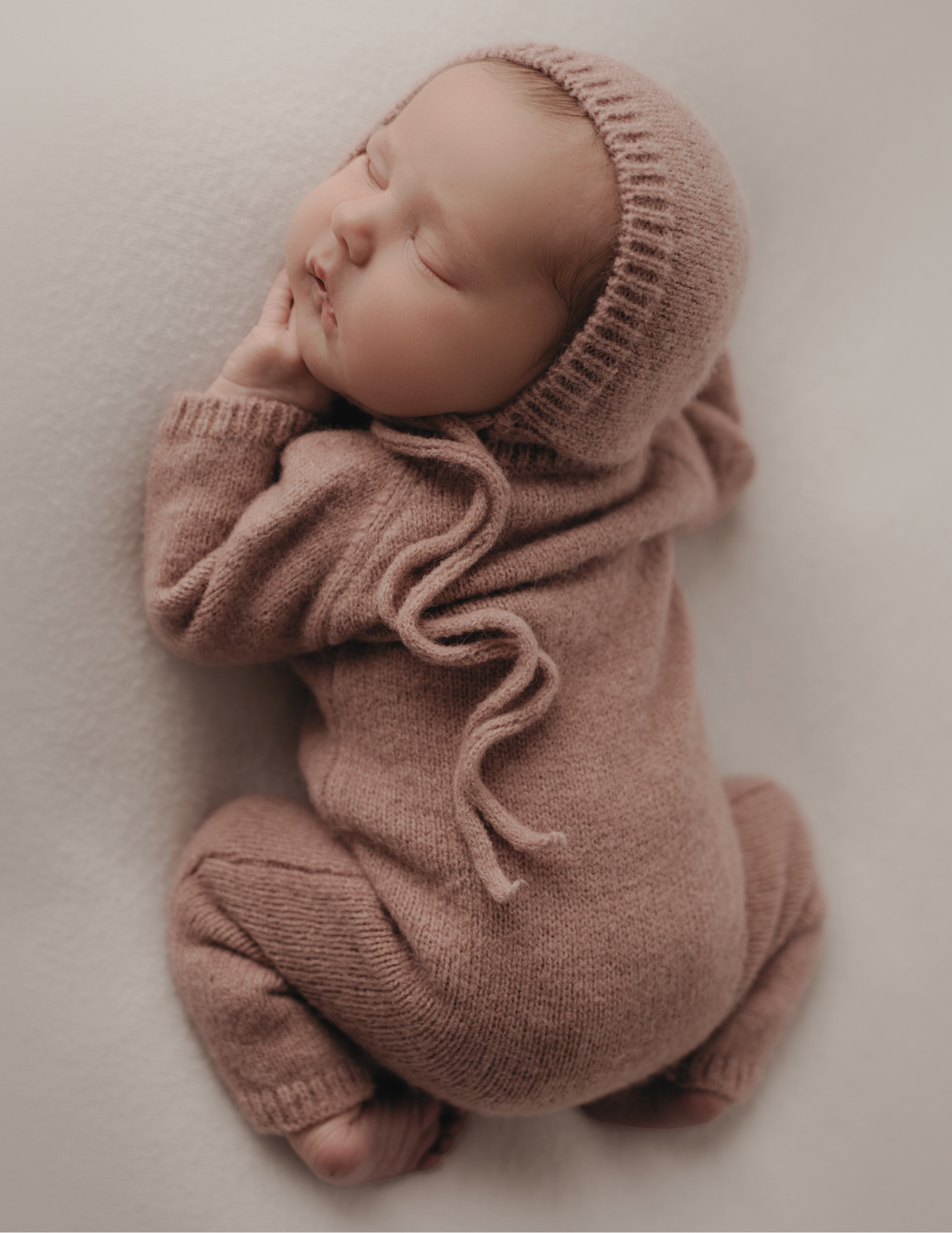 Peaceful sleeping newborn baby in dusty pink knit romper and matching bonnet on white background.