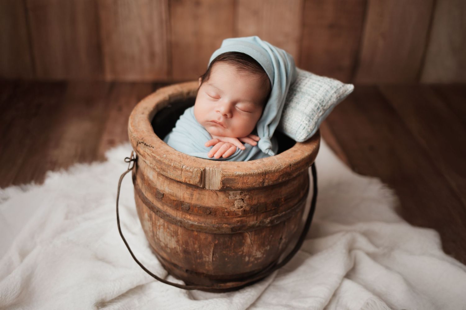 Peaceful newborn baby in blue wrap sleeping inside a rustic wooden bucket with pillow.