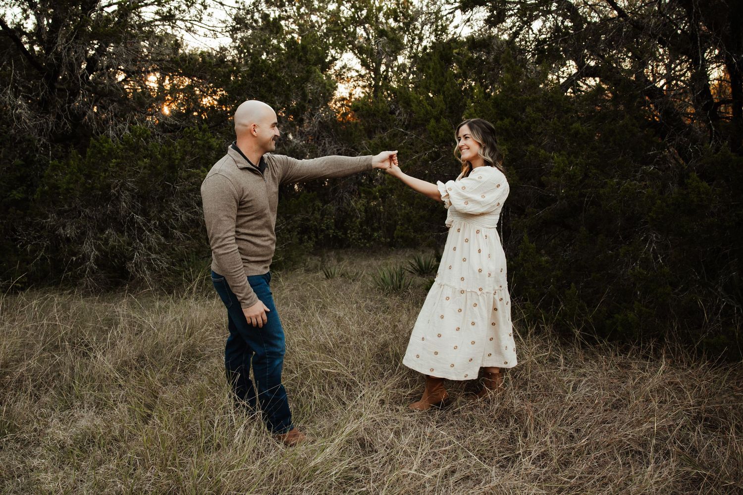 Couple dancing and holding hands in a golden sunset field surrounded by trees.