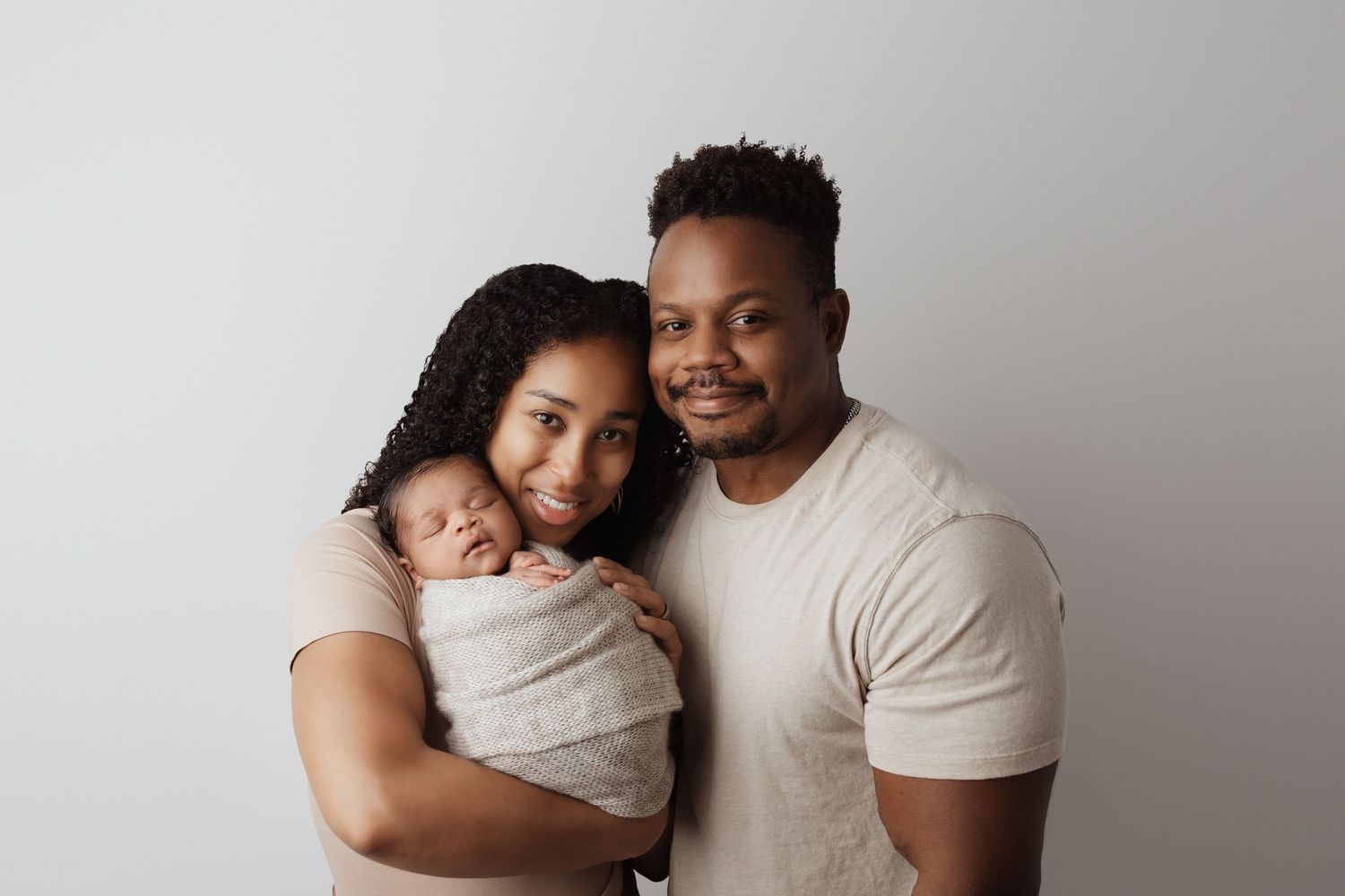 A happy couple poses together holding their swaddled newborn baby against a clean white studio backdrop.