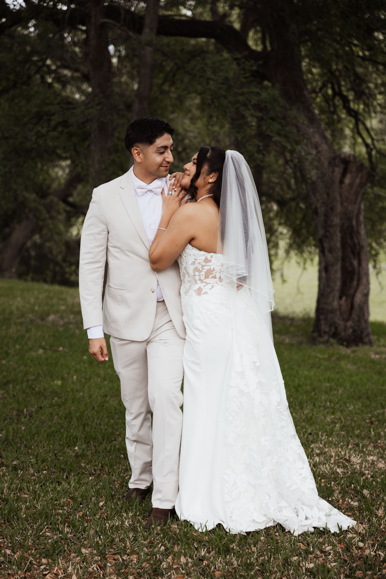 A bride and groom share a romantic moment outdoors, with the bride in a lace gown and veil, groom in a tan suit.