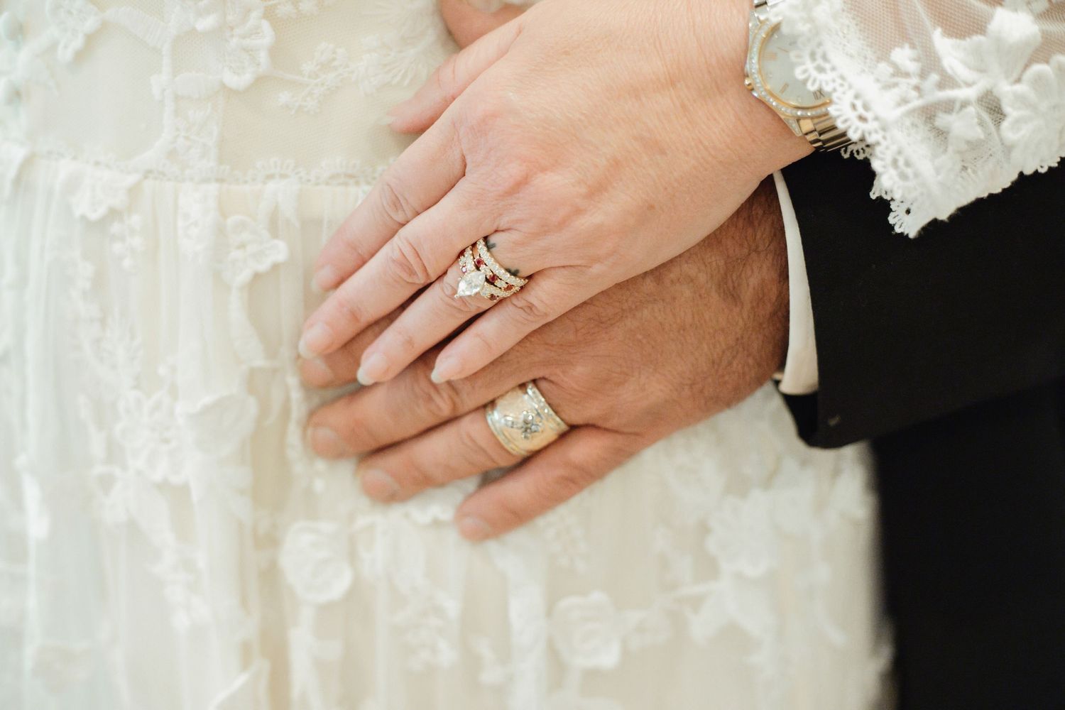 Close-up of newlyweds hands showing wedding rings, bride in lace dress and groom in black suit.