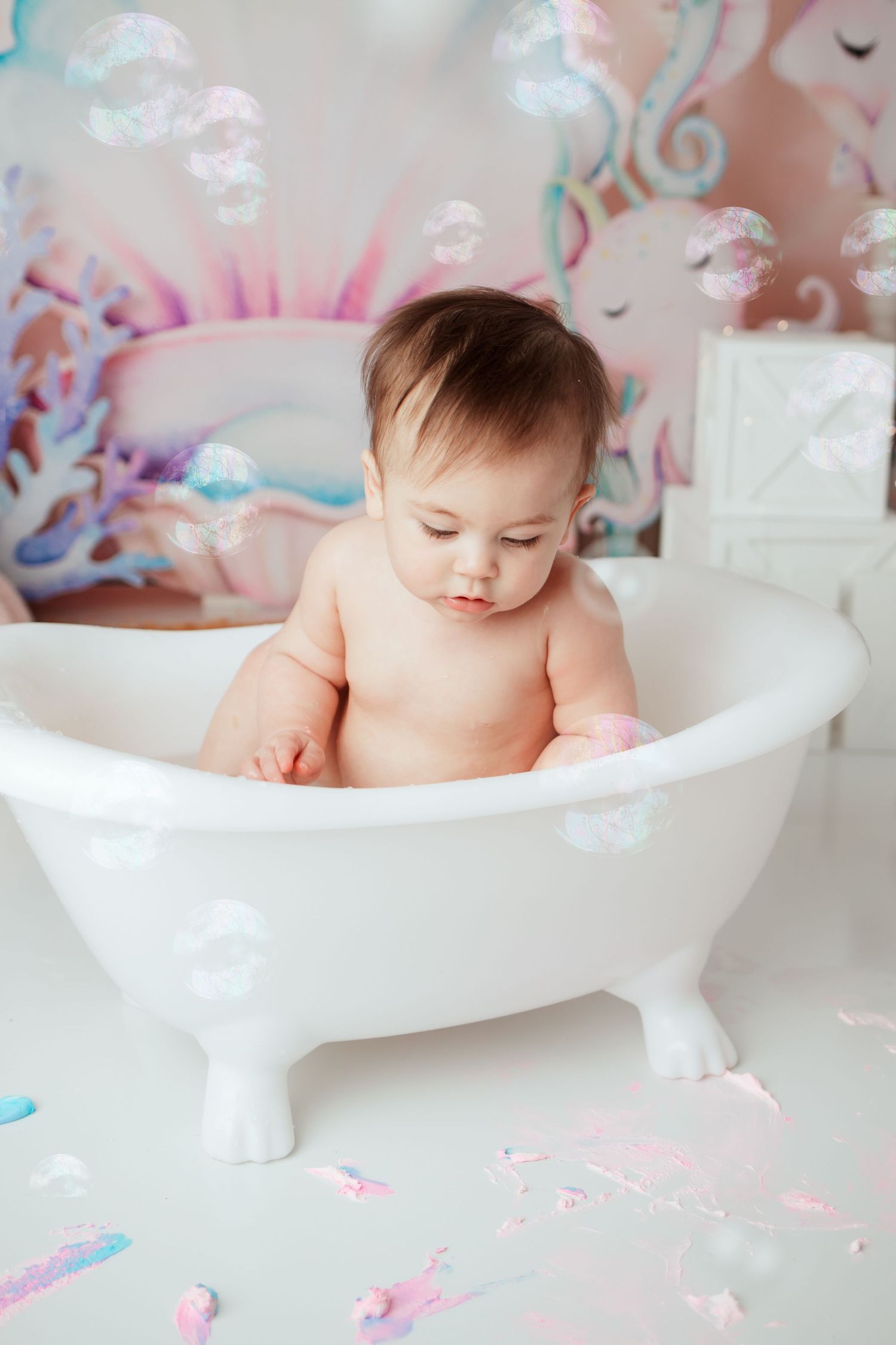 Baby sitting in white clawfoot bathtub during cake smash photo session with unicorn backdrop.