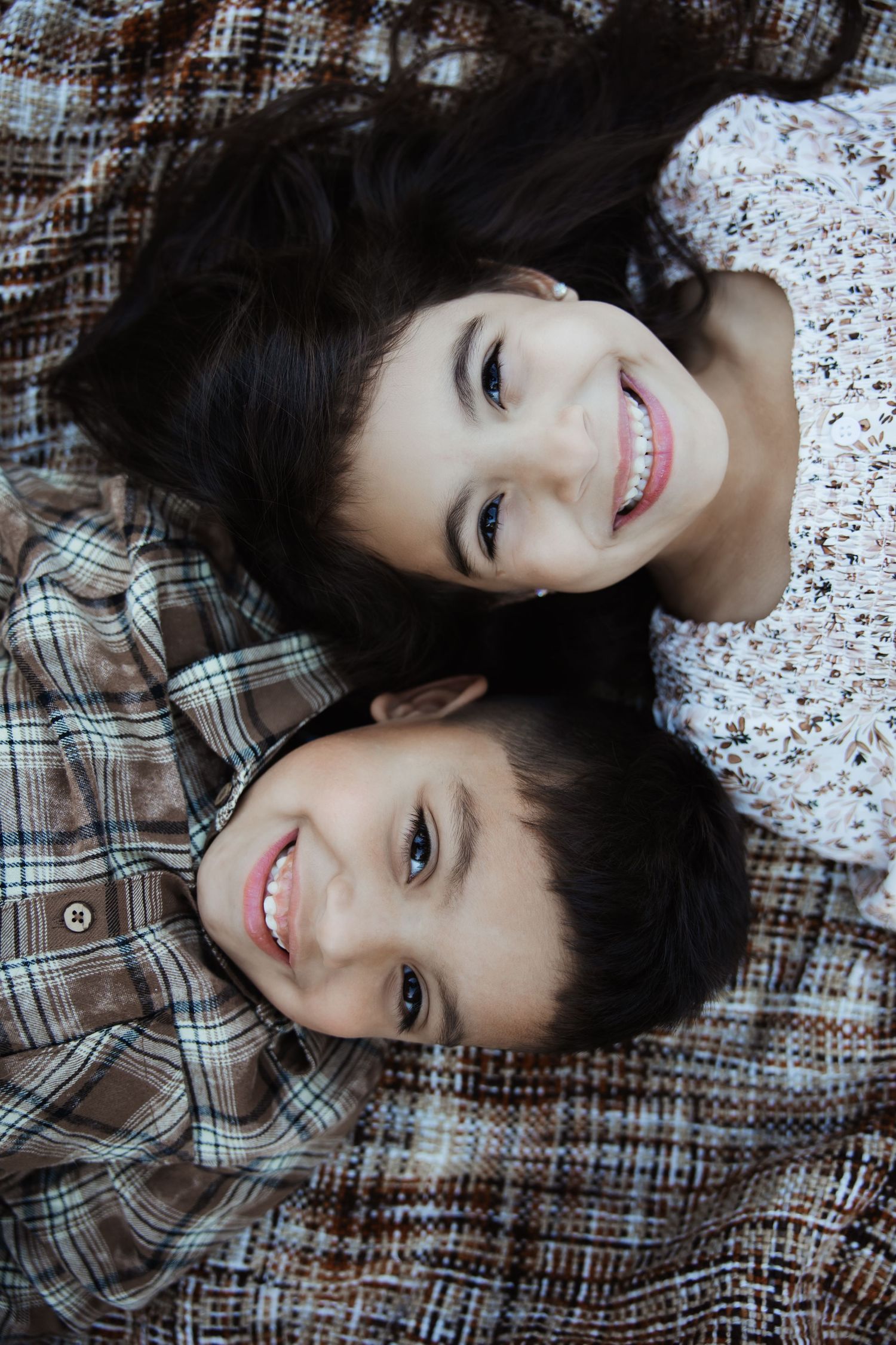 Two smiling siblings lying on a plaid blanket looking up at the camera.