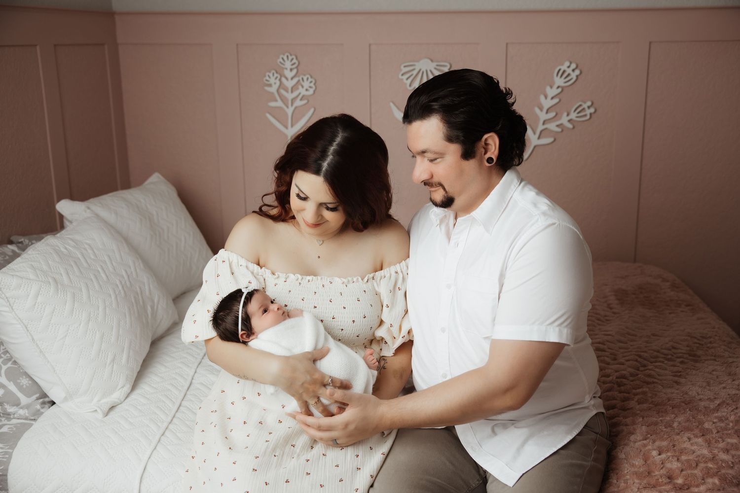 New parents sitting on bed holding swaddled newborn baby in beautifully decorated pink nursery.
