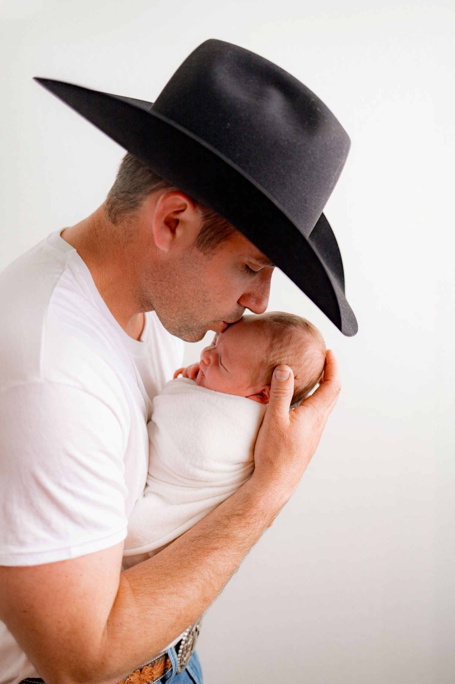 Father in black cowboy hat and white shirt gently kissing his sleeping newborn baby.