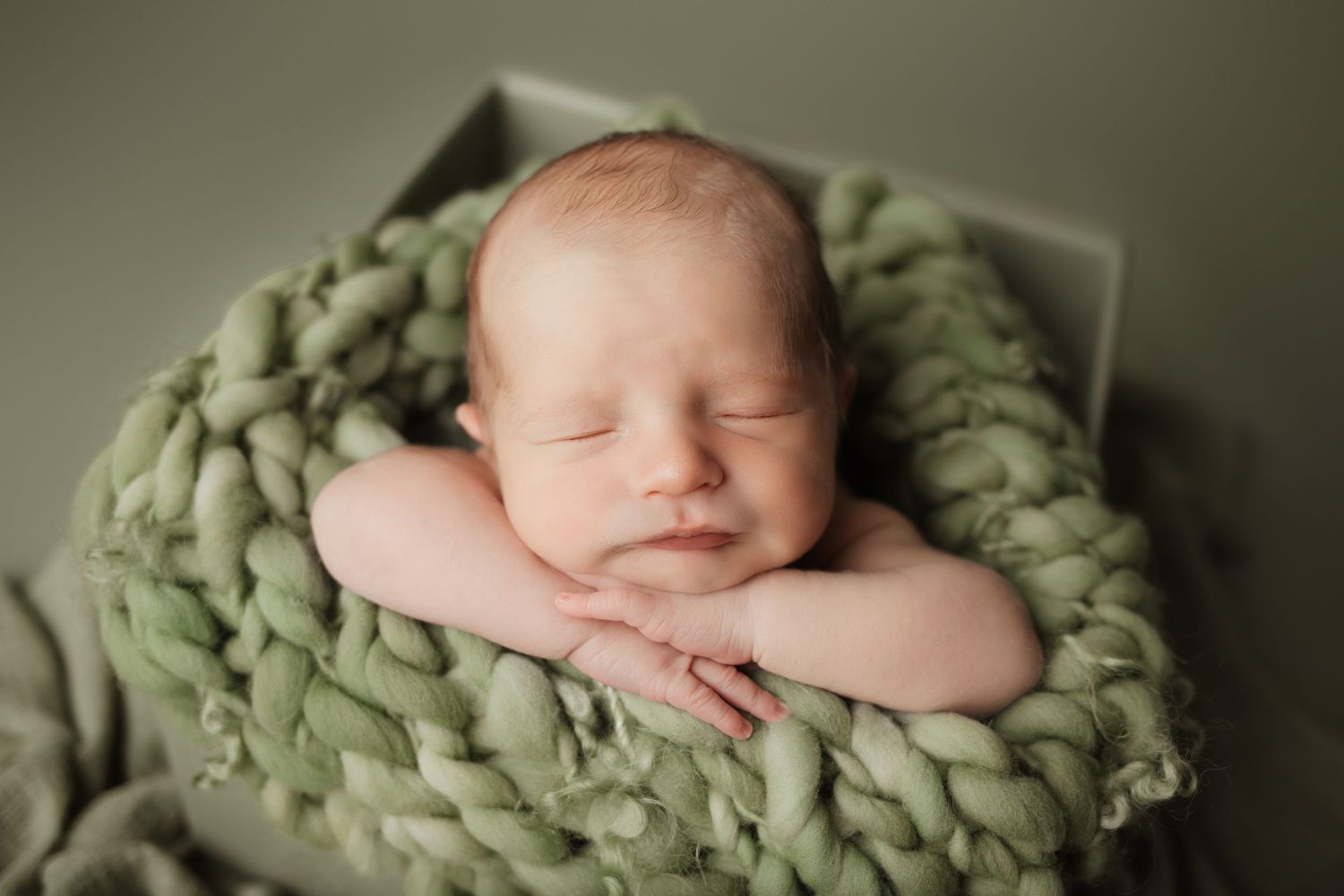 Peaceful newborn baby sleeping on a chunky green knit blanket in a soft sage-colored bowl prop.