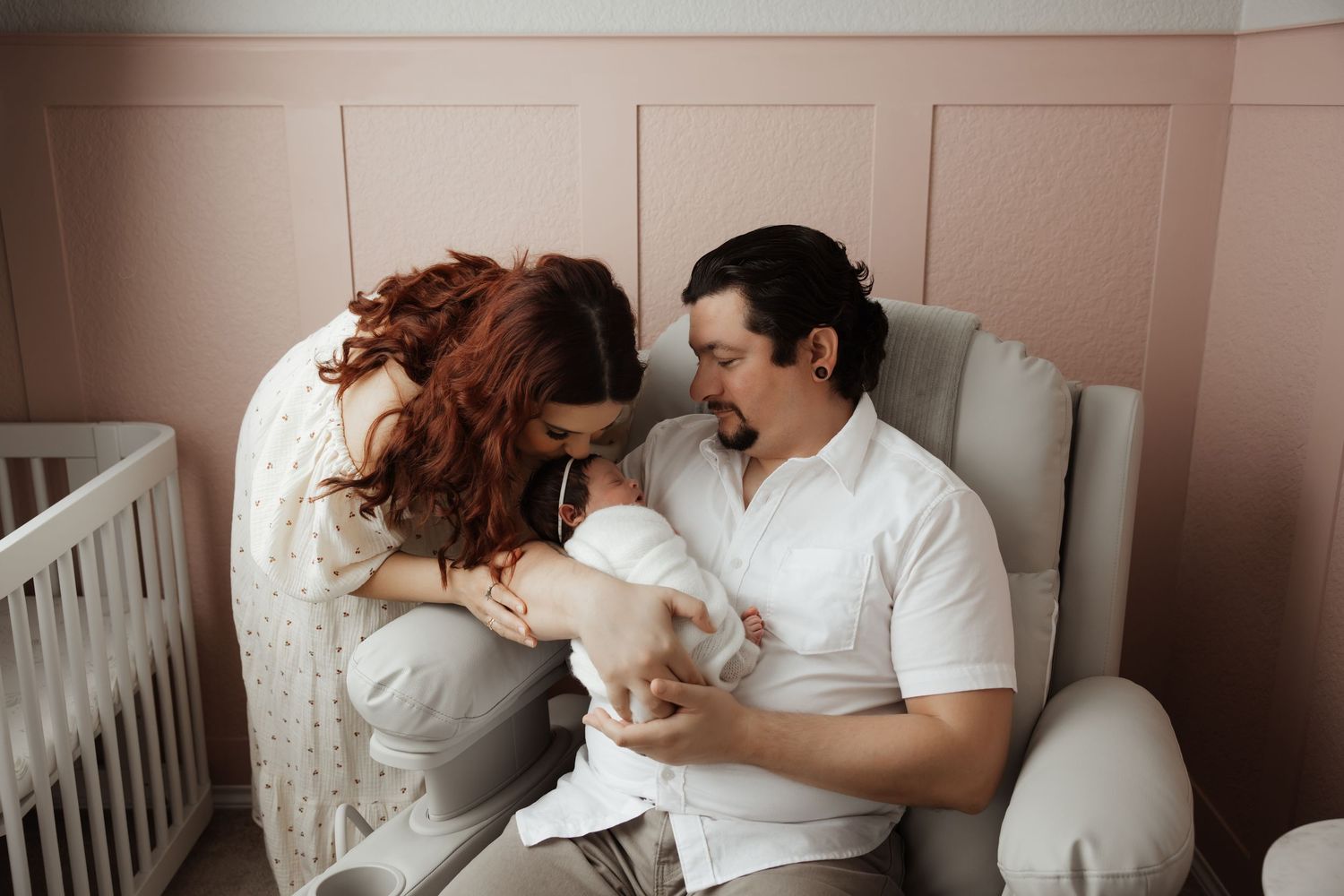 New parents sitting in nursery chair admiring their swaddled newborn baby in pink room.