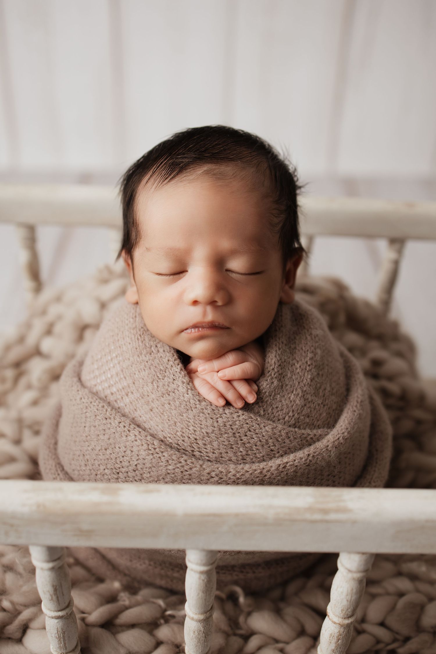 Swaddled newborn with dark hair sleeping upright in brown knit wrap inside white wooden crib.