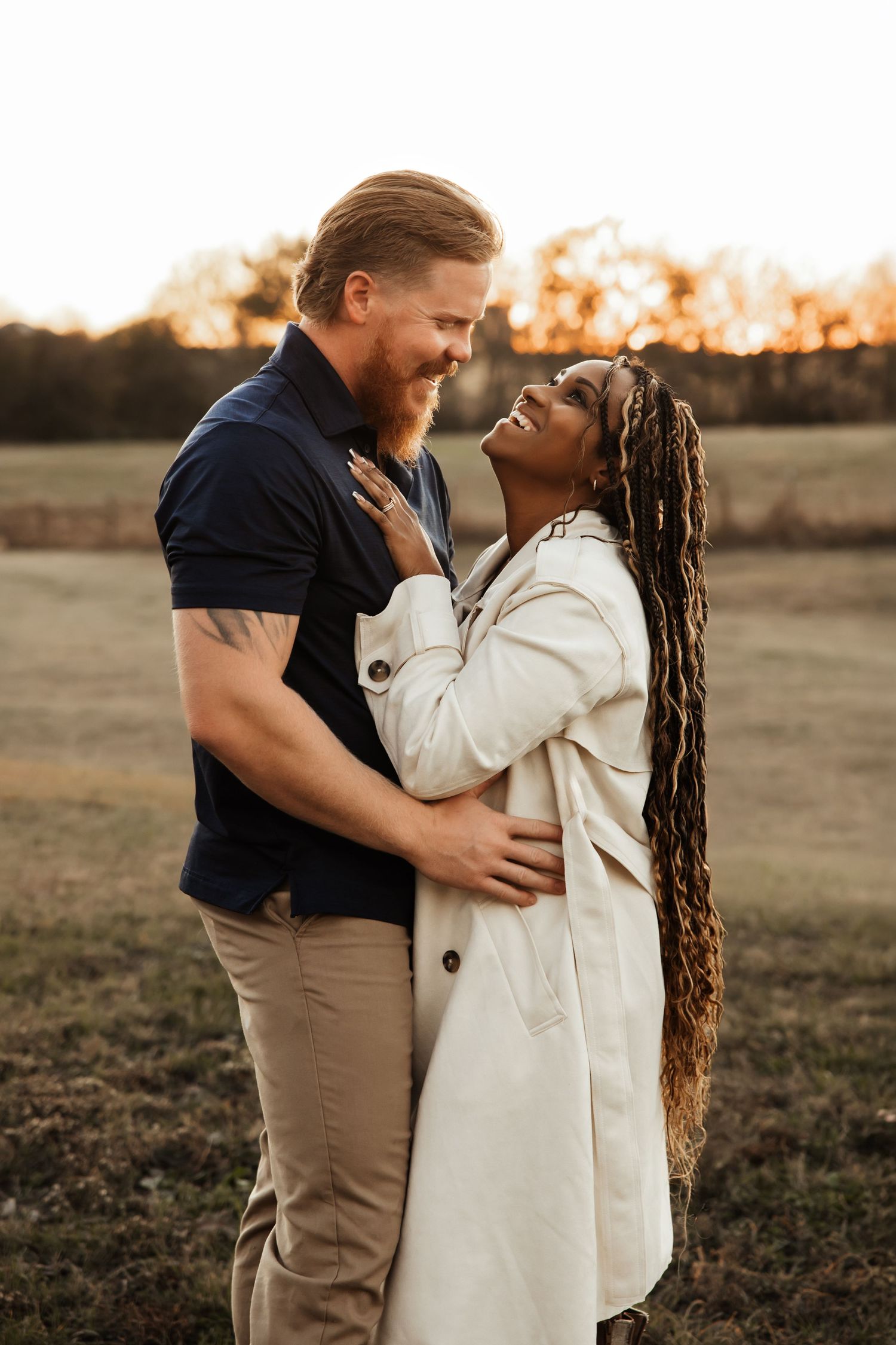 An interracial couple embraces and smiles at each other during a golden hour outdoor shoot.