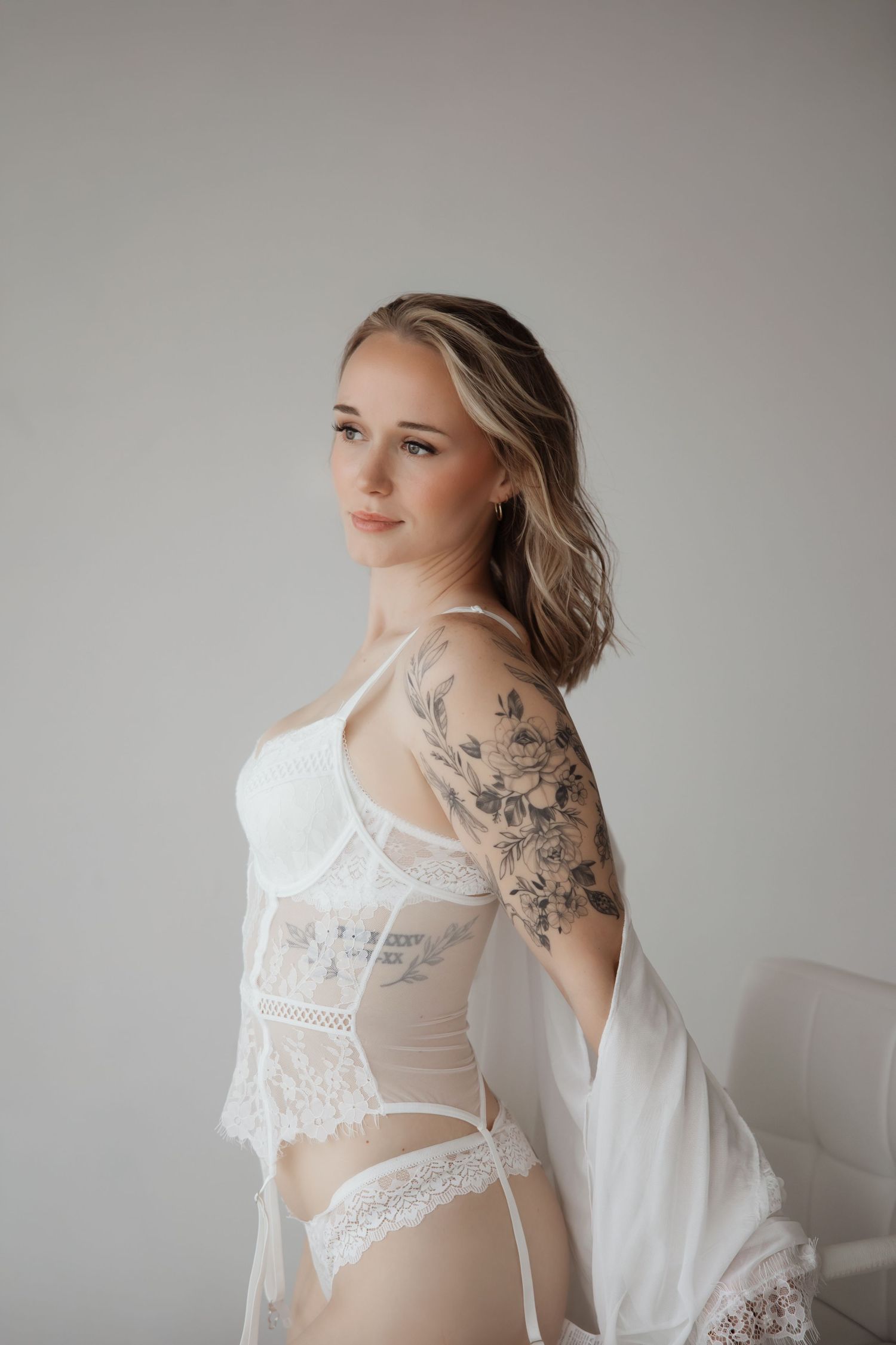 Young woman in white lace lingerie and sheer robe, showcasing floral arm tattoo, posing against light gray background.