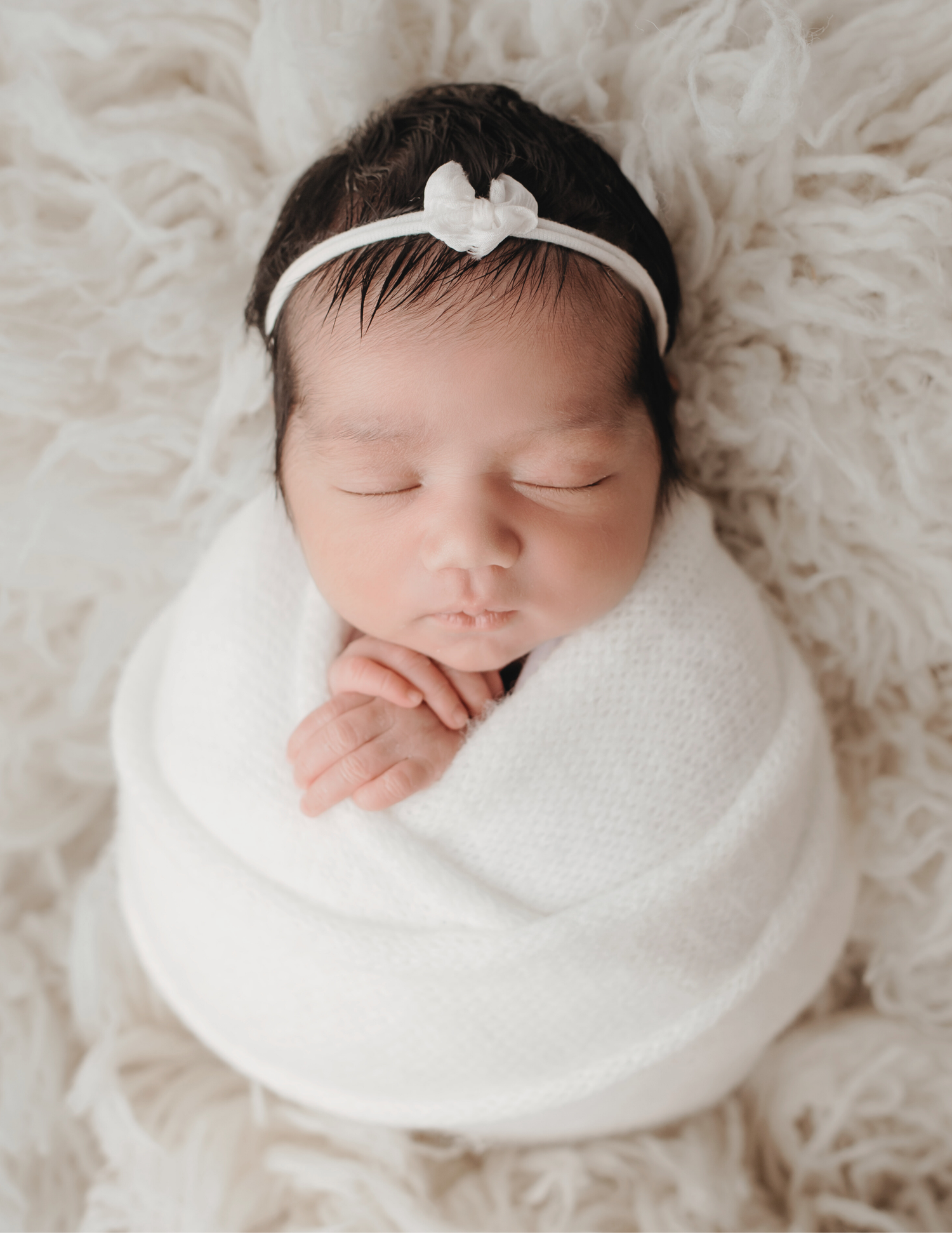 Sleeping newborn baby girl wrapped in white swaddle with a delicate bow headband on fluffy backdrop.