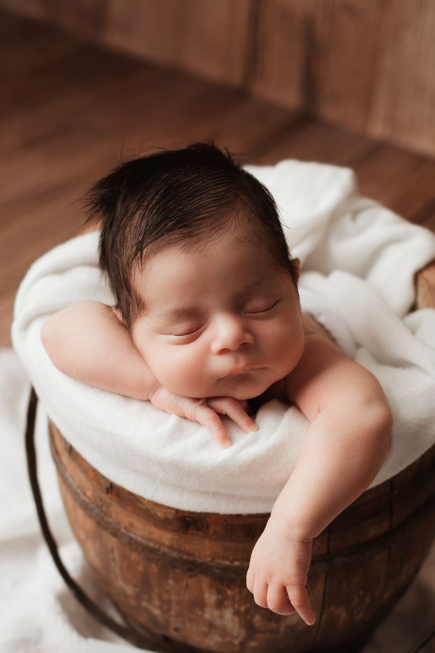 Sleeping newborn with dark hair resting peacefully in a rustic wooden barrel with white blanket.