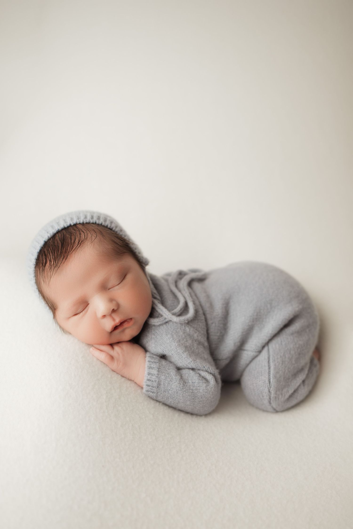 Sleeping newborn baby in gray knit romper and hat resting on a cream blanket.