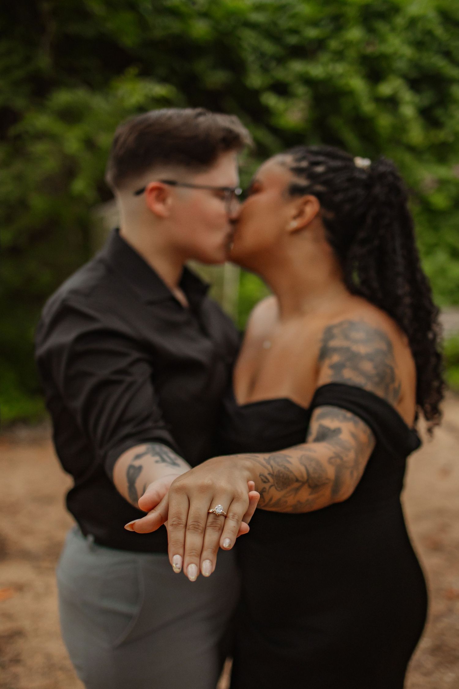 Couple kissing outdoors displaying engagement ring with tattoos visible, wearing black outfits against green foliage backdrop.