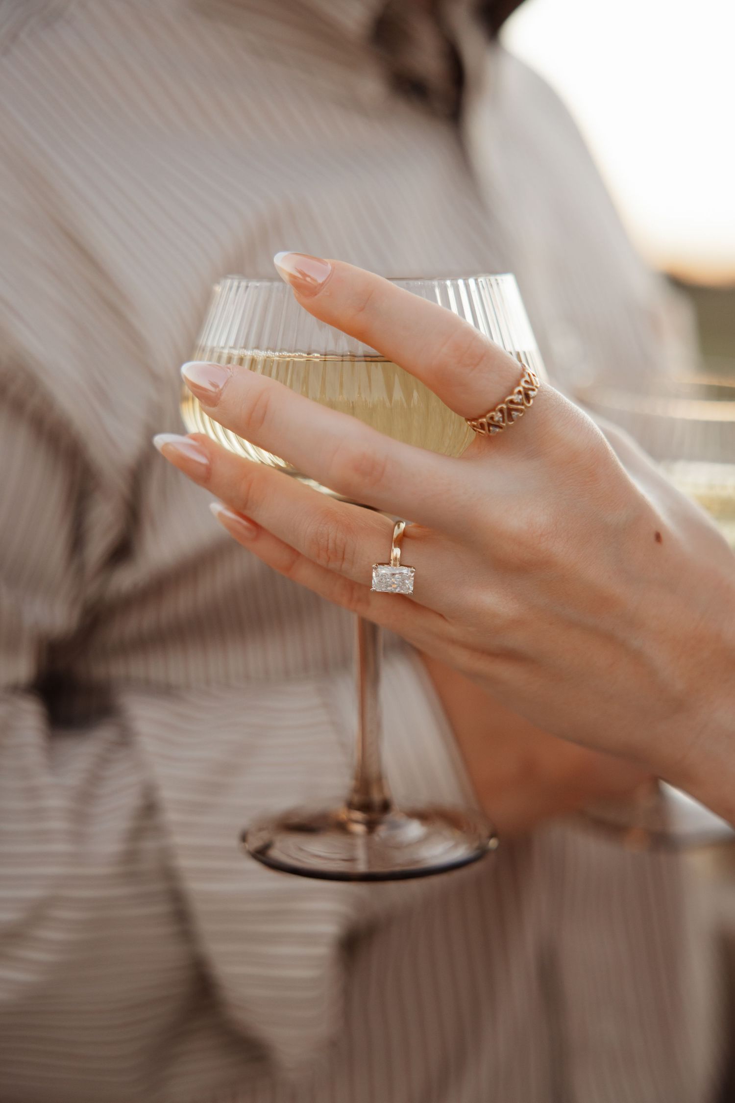Woman holding ribbed glass of white wine wearing elegant emerald-cut diamond ring and gold band on manicured hand.