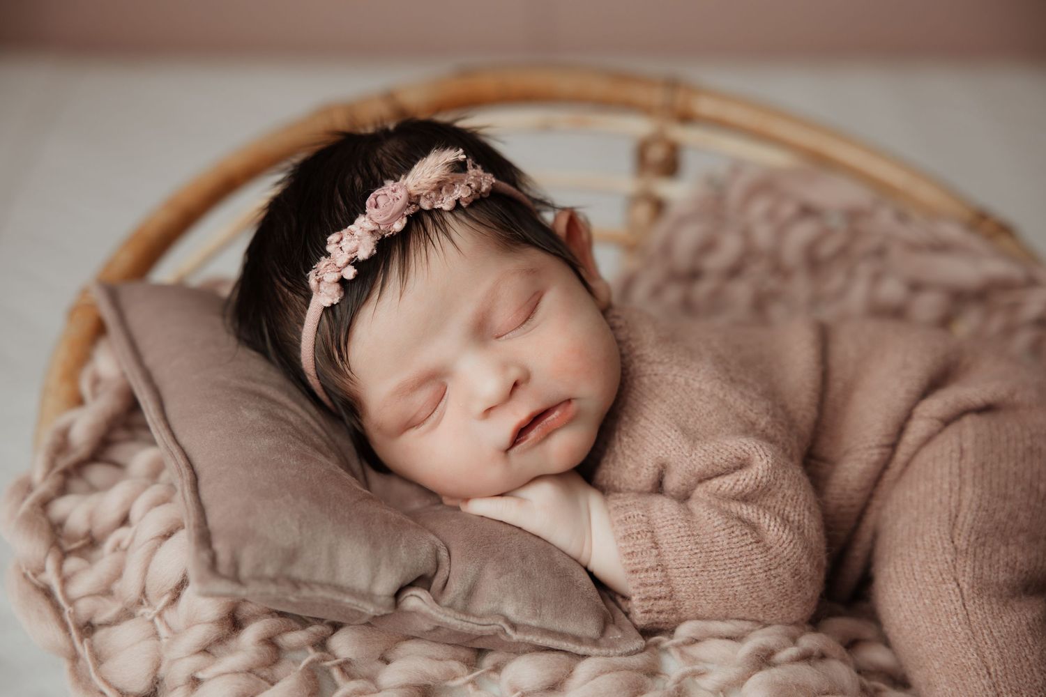 Close-up of sleeping newborn baby girl wearing pink floral headband, dressed in a soft pink knit romper.