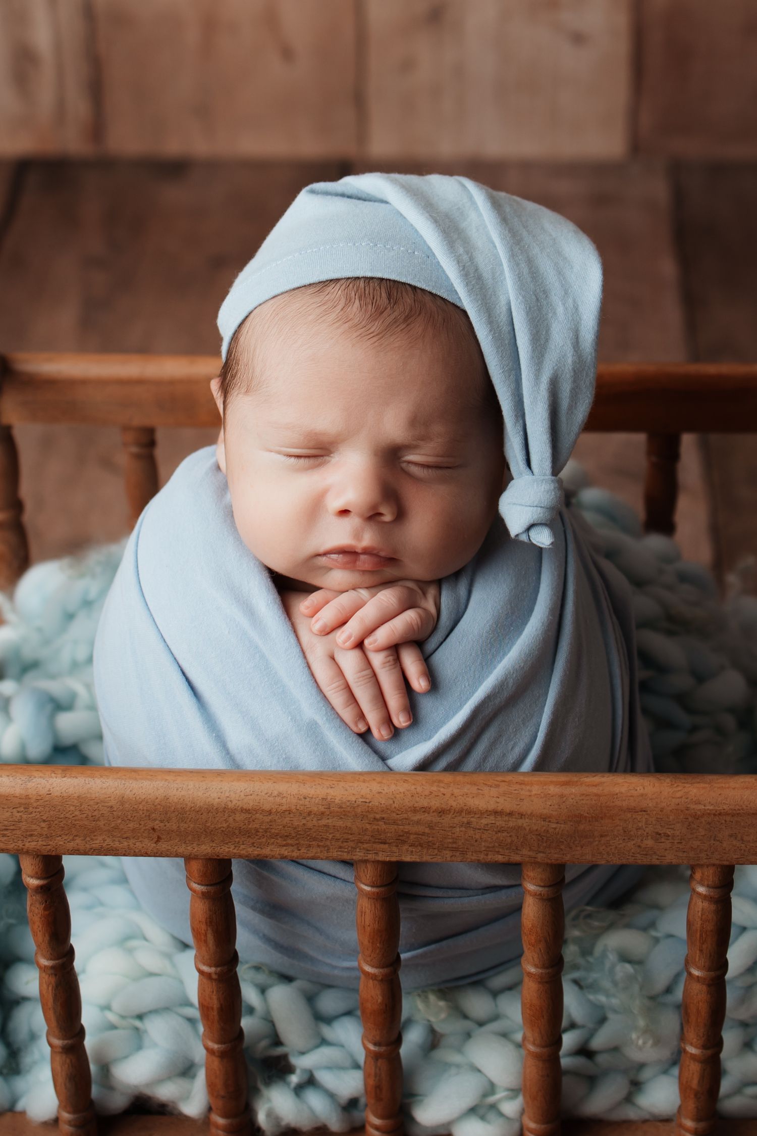 Peaceful newborn baby swaddled in blue wrap wearing a knotted sleep cap in a wooden crib.