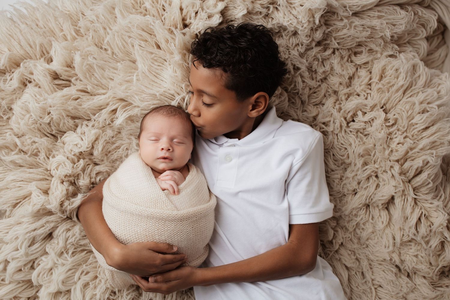 Older brother gently kissing swaddled newborn baby sibling on a fluffy beige fur rug.