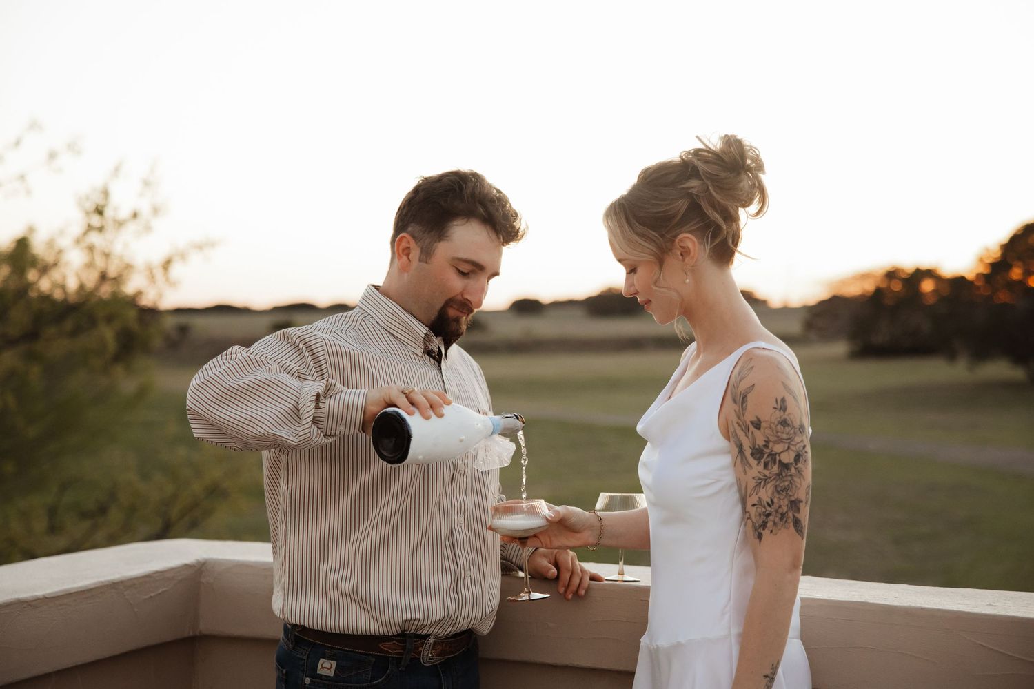 Groom pouring champagne for tattooed bride at sunset on an outdoor terrace during their wedding celebration.