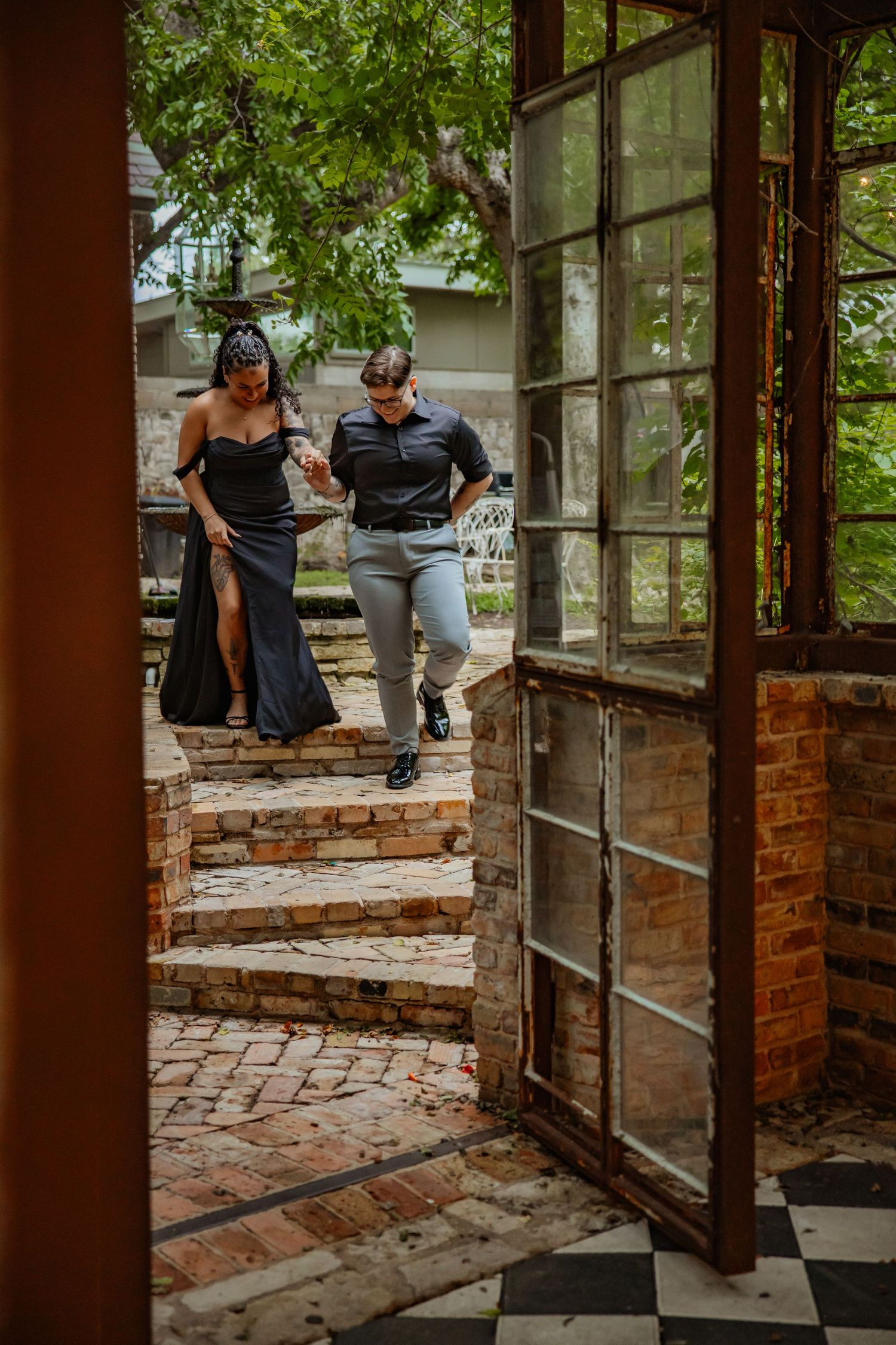 Couple in formal attire walking down brick steps through rustic iron-framed glass doors at an elegant outdoor venue.