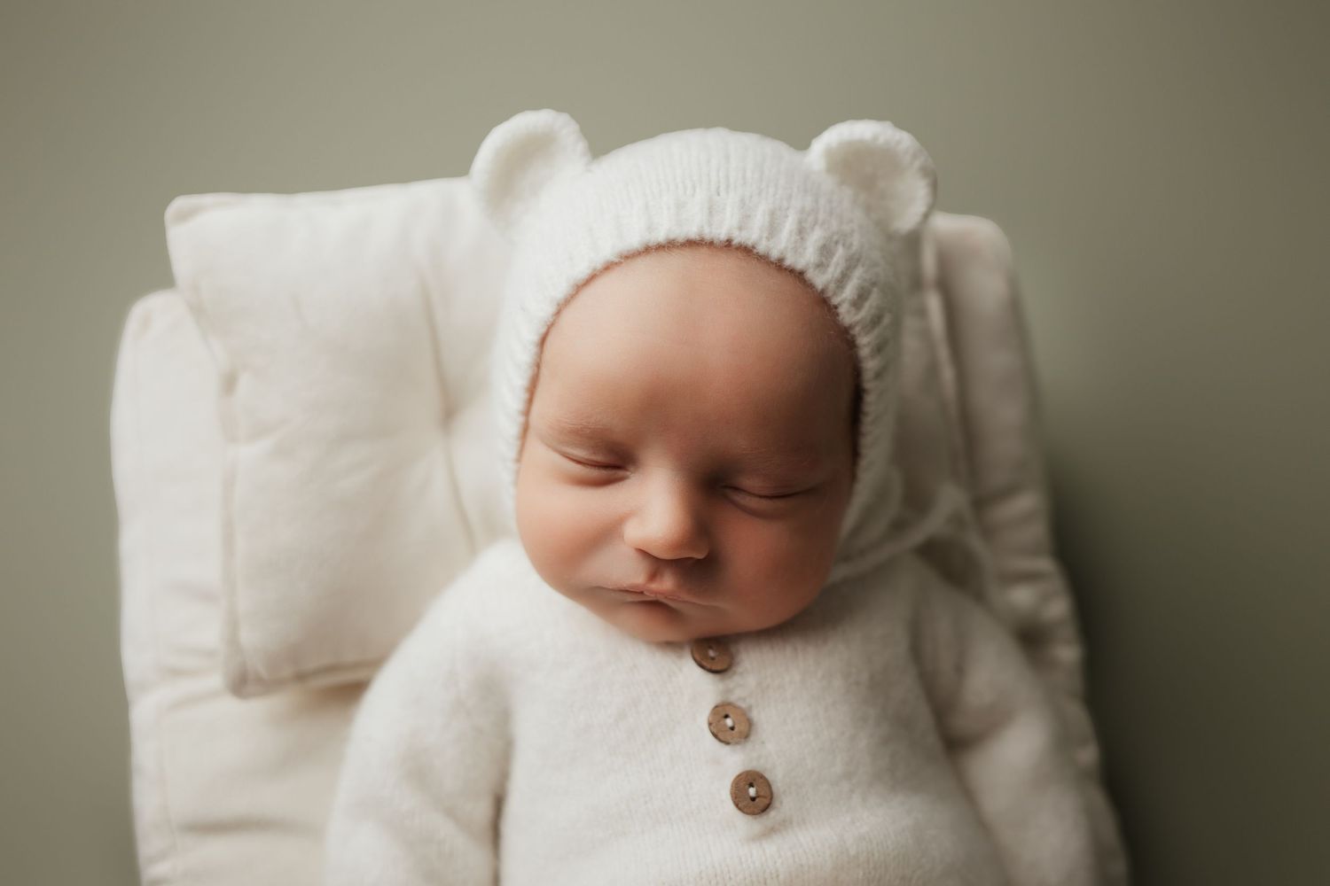 Peaceful newborn baby in white knit bear bonnet and romper rests on white cushioned chair.