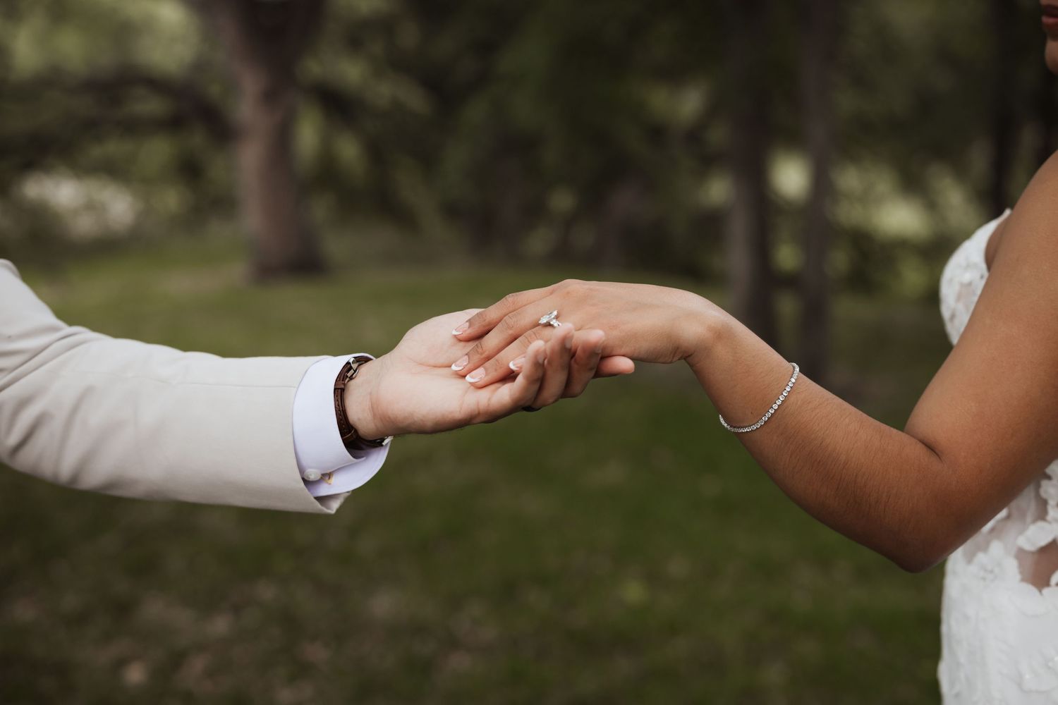 Bride and groom holding hands outdoors, showcasing wedding rings and bracelets in a romantic garden ceremony.