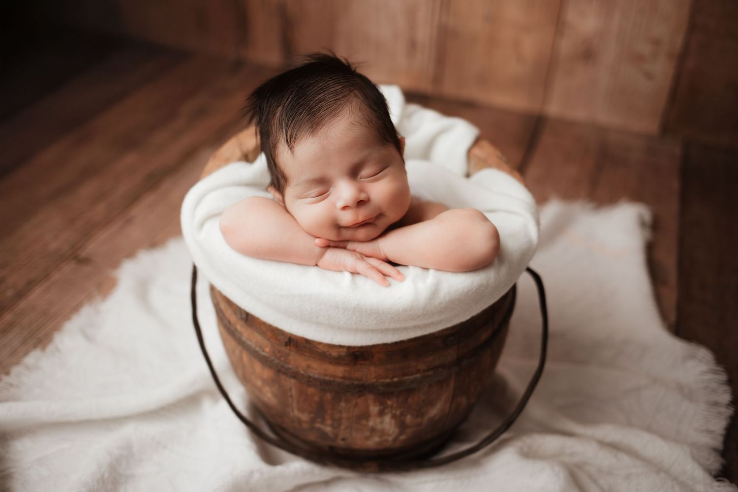 Smiling newborn baby sleeping in a rustic wooden bucket with white linen lining.