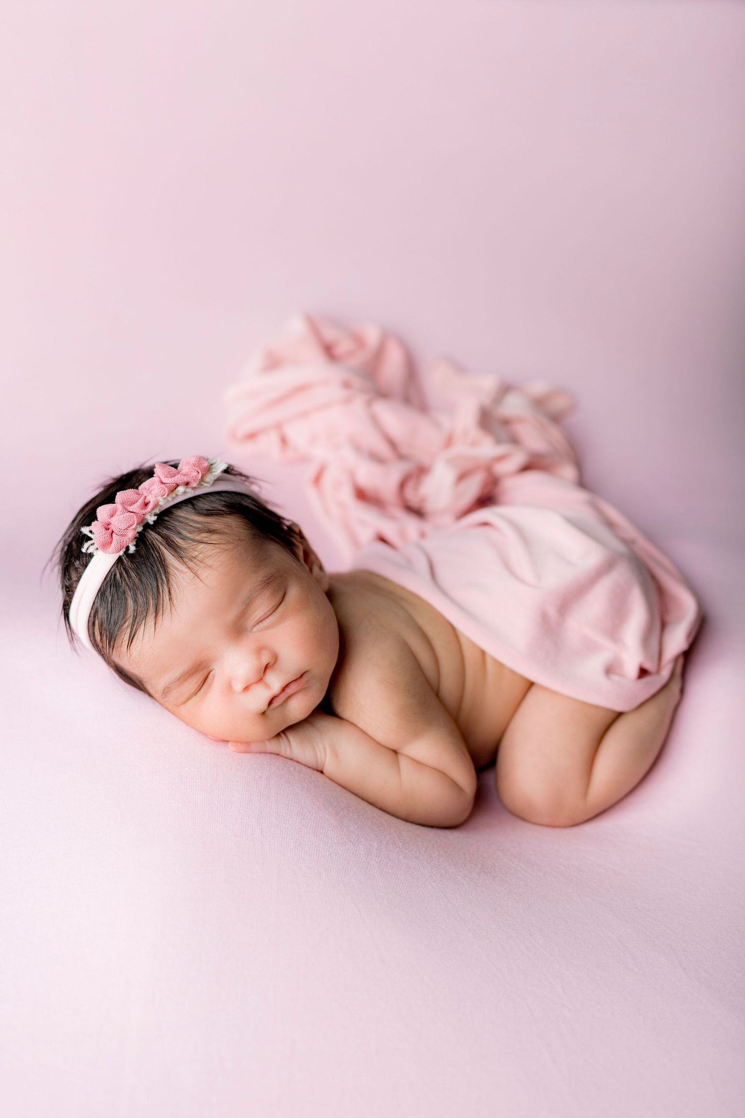 Sleeping newborn baby girl with pink bow headband posed on soft pink blanket with pink wrap.