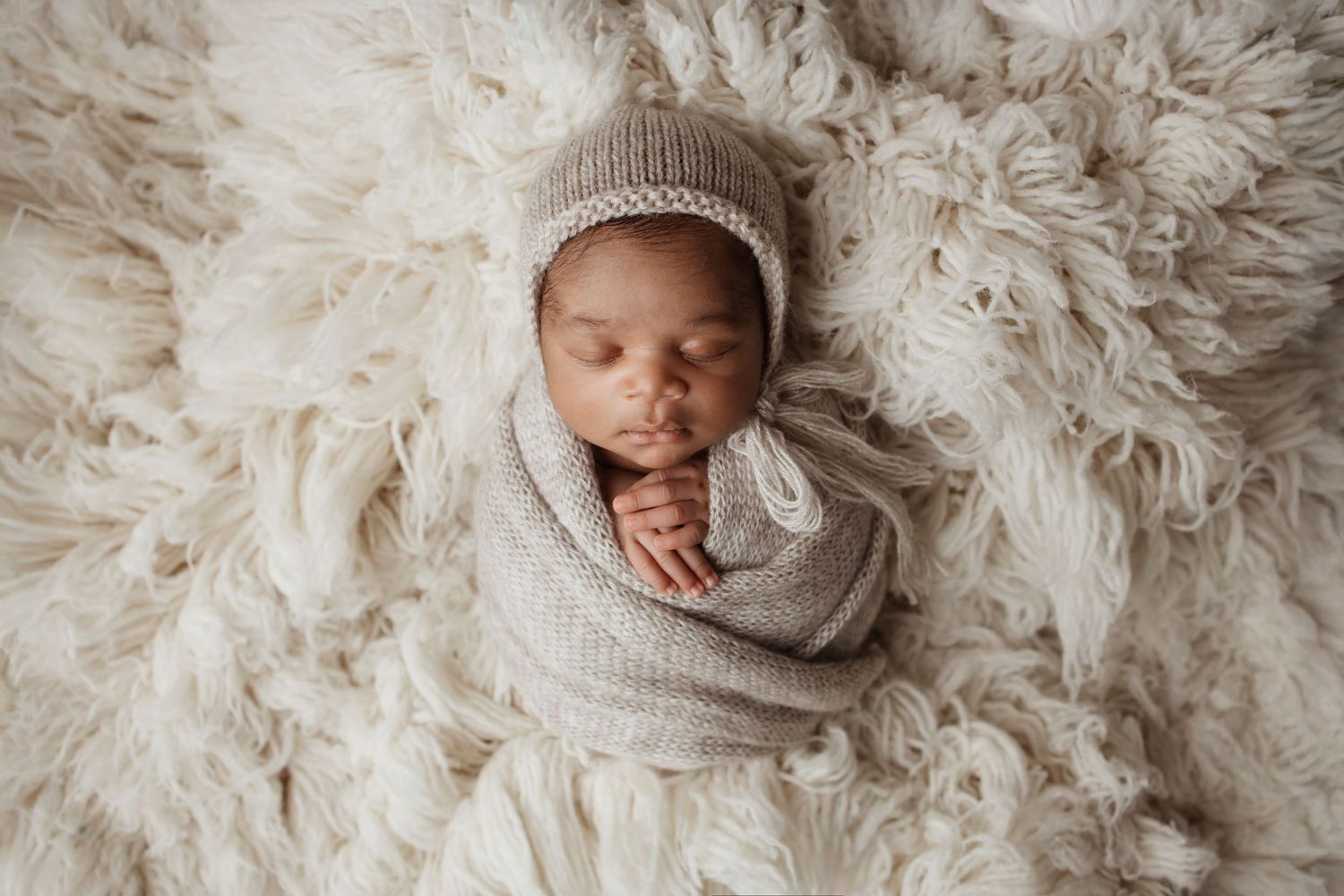 A swaddled newborn baby wearing a knit bonnet sleeps peacefully on a fluffy cream fur rug.
