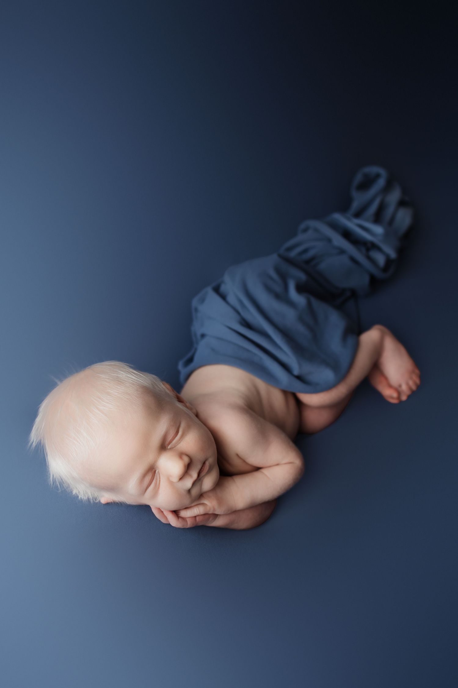 Newborn baby with blonde hair sleeping on a blue background wrapped in a matching blue cloth.